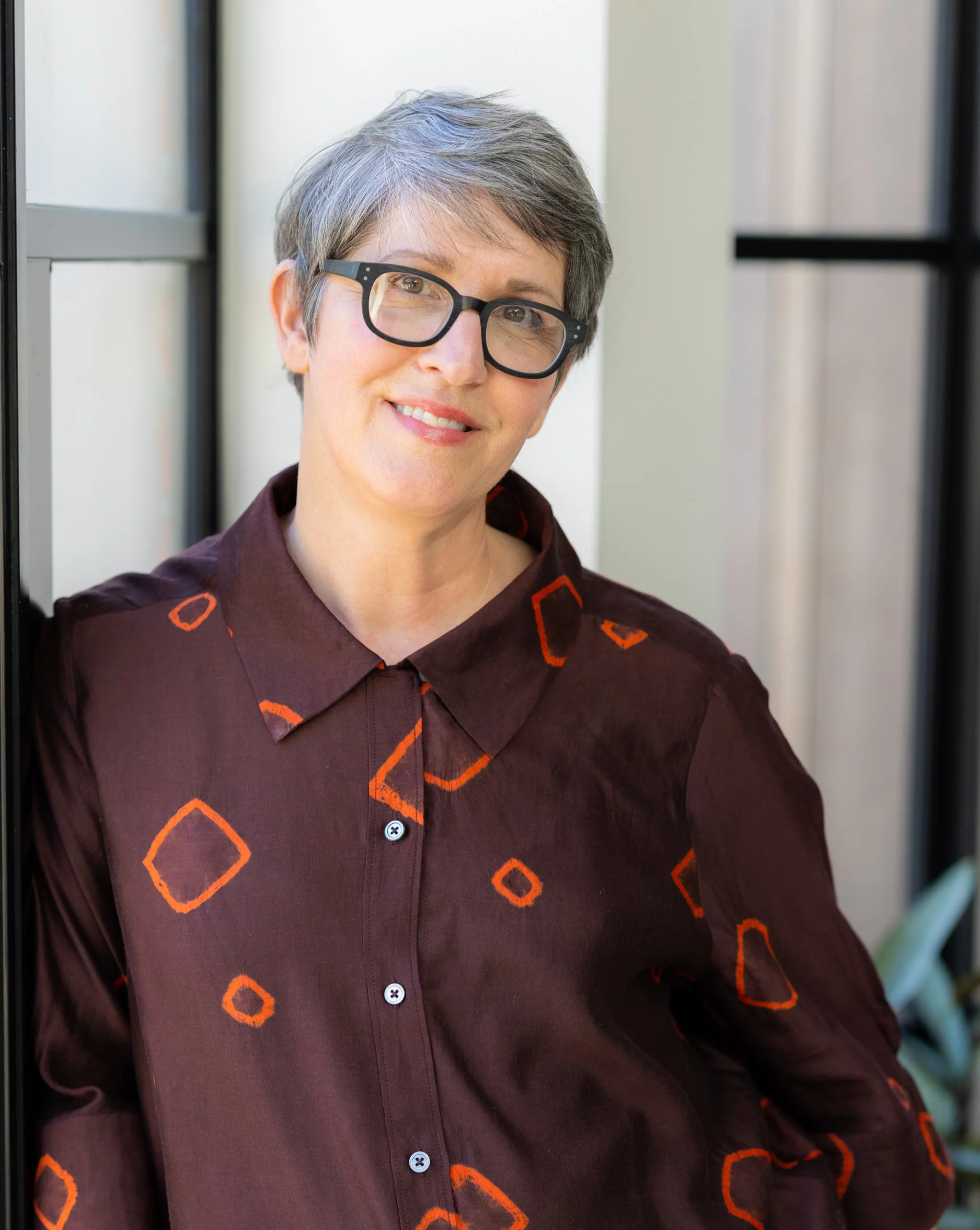 A woman with short gray hair and black glasses smiling, wearing a dark brown shirt with orange geometric patterns, sitting near a window.