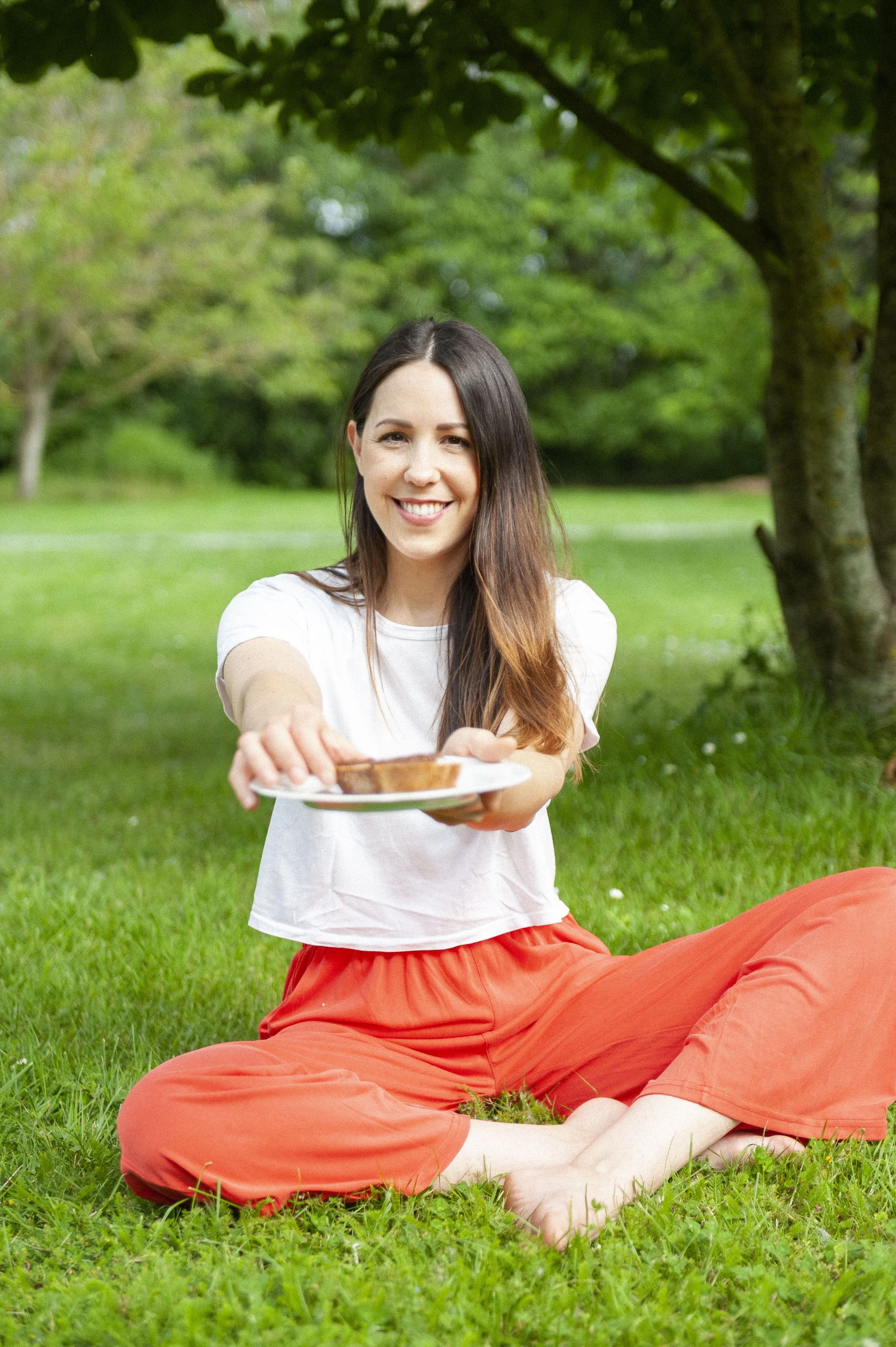 Femme assise dans l'herbe, souriant, tenant une assiette avec une collation, arbres en arrière-plan.