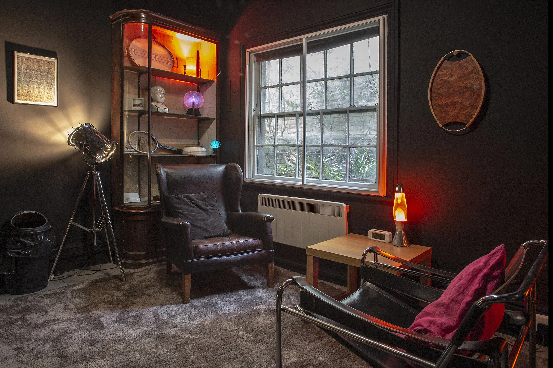 The Den, a cosy dark therapy room with 2 chairs and table with a window into a garden