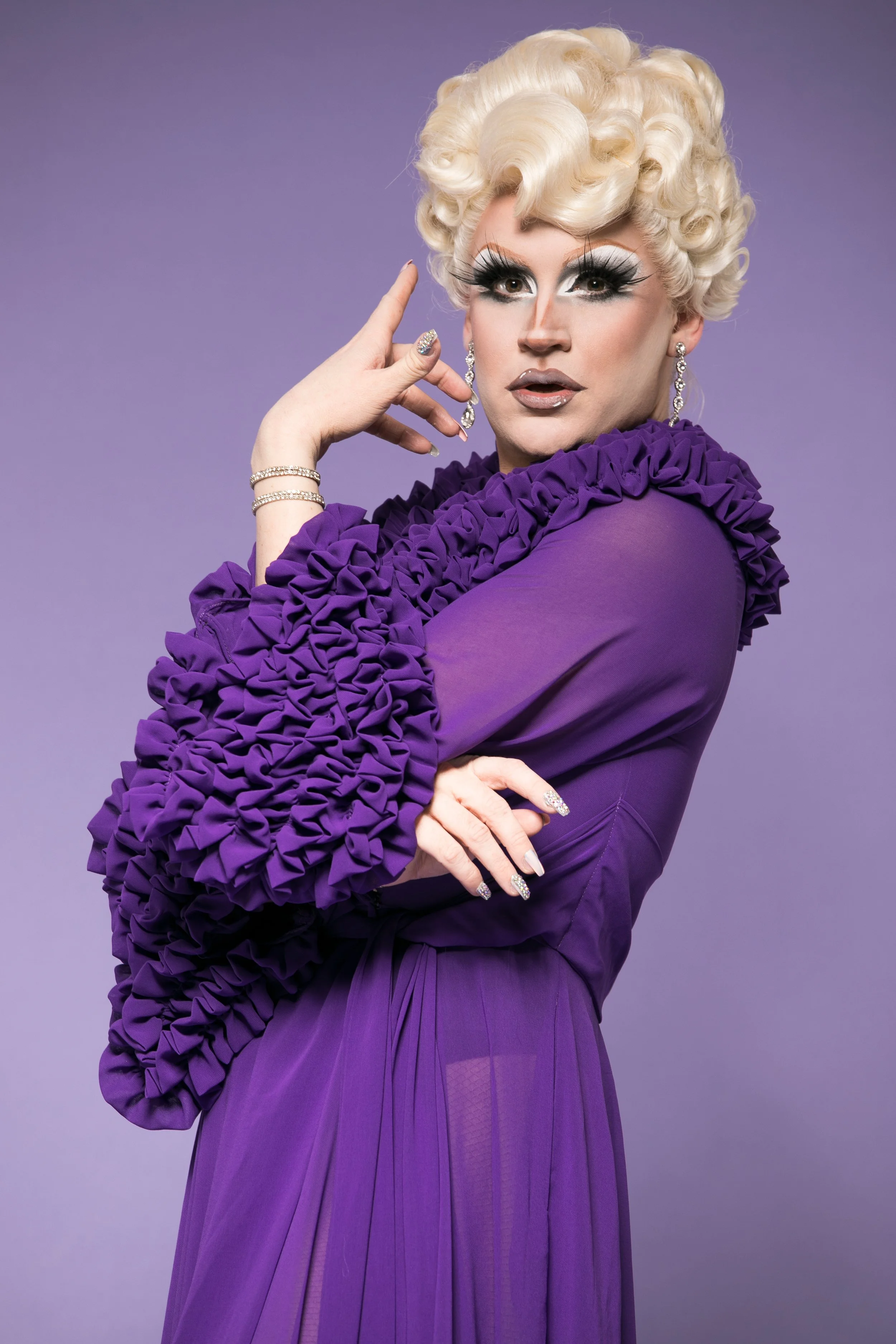 Person with curly blonde hair wearing a purple dress with ruffled sleeves, large earrings, and dramatic makeup, posing against a purple background.