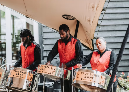 Three steel drum musicians wearing black shirts and red vests performing outdoors.