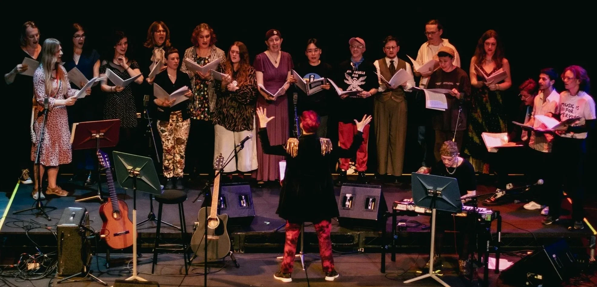 A choir of diverse people singing on stage, directed by a conductor, with musical instruments and equipment in front.