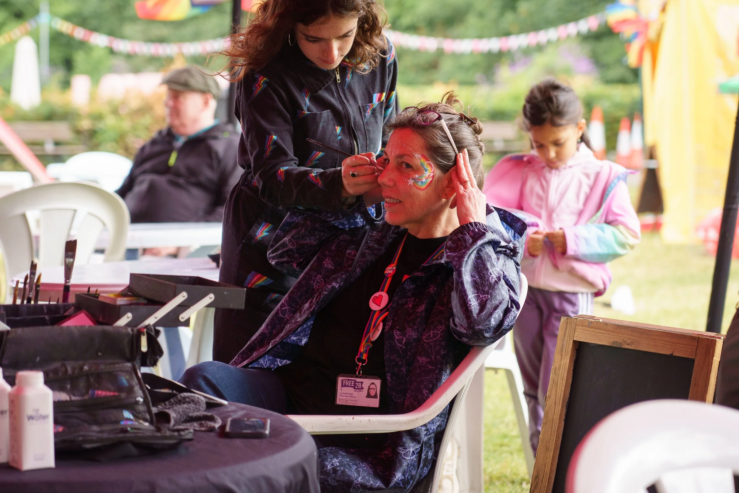 A woman sitting at a table getting her face painted with a colorful rainbow design at an outdoor festival or fair, with a young girl and other people in the background.
