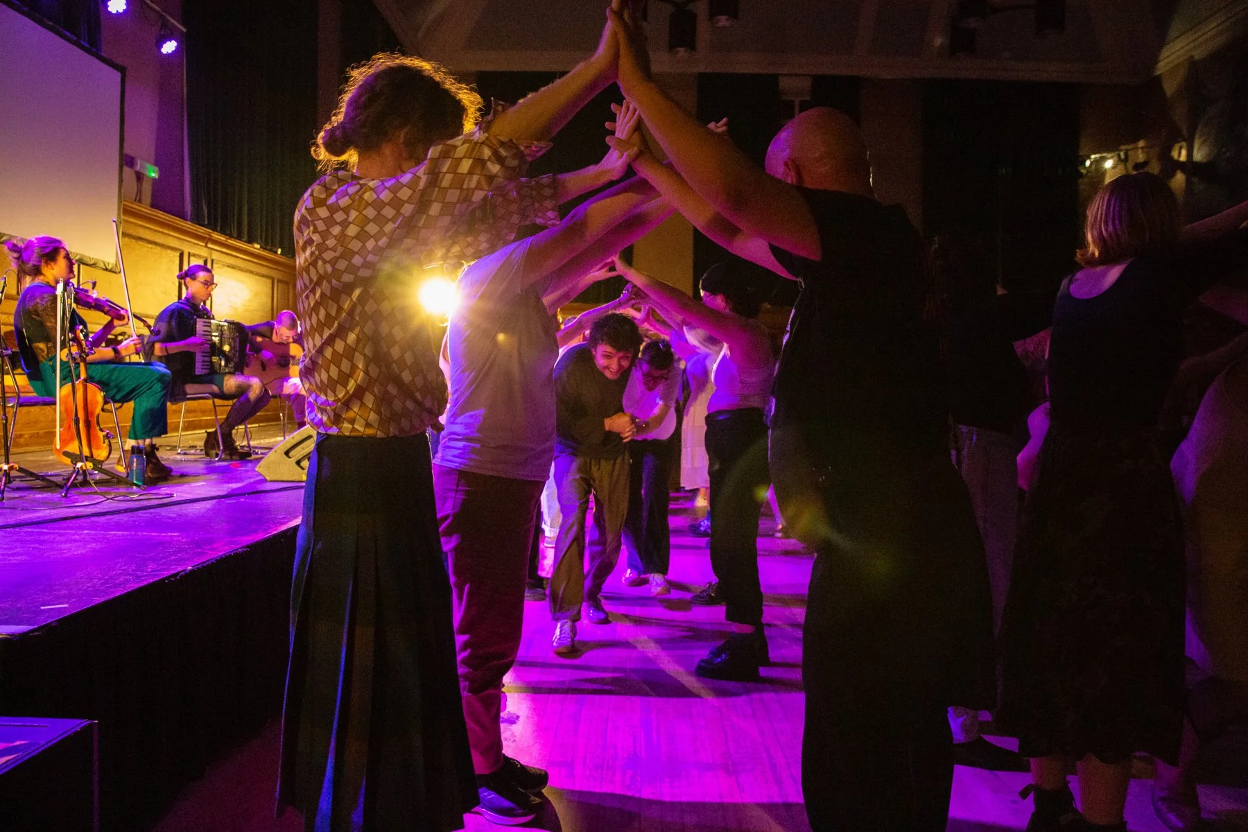 People dancing in a circle at a concert or event, with a band playing on stage in the background, under purple stage lighting.