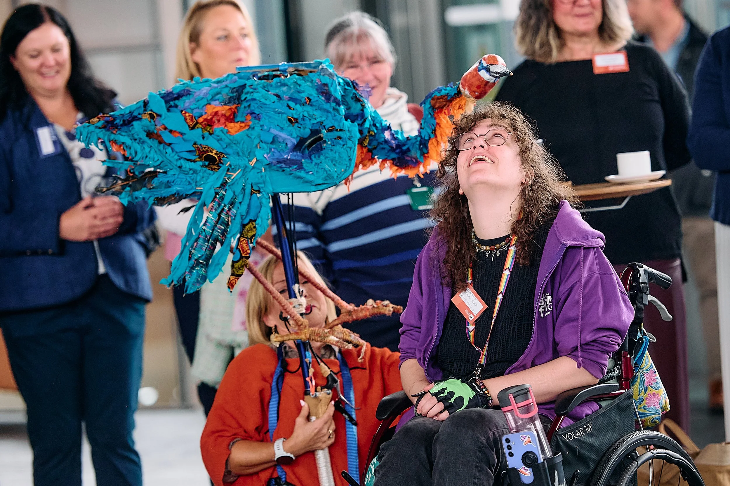 A group of smiling women, some wearing name tags, gathered around a colorful puppet resembling a bird or dinosaur with blue, orange, and patterned fabric. One woman in a purple jacket in a wheelchair interacts with the puppet, while others look on and smile.