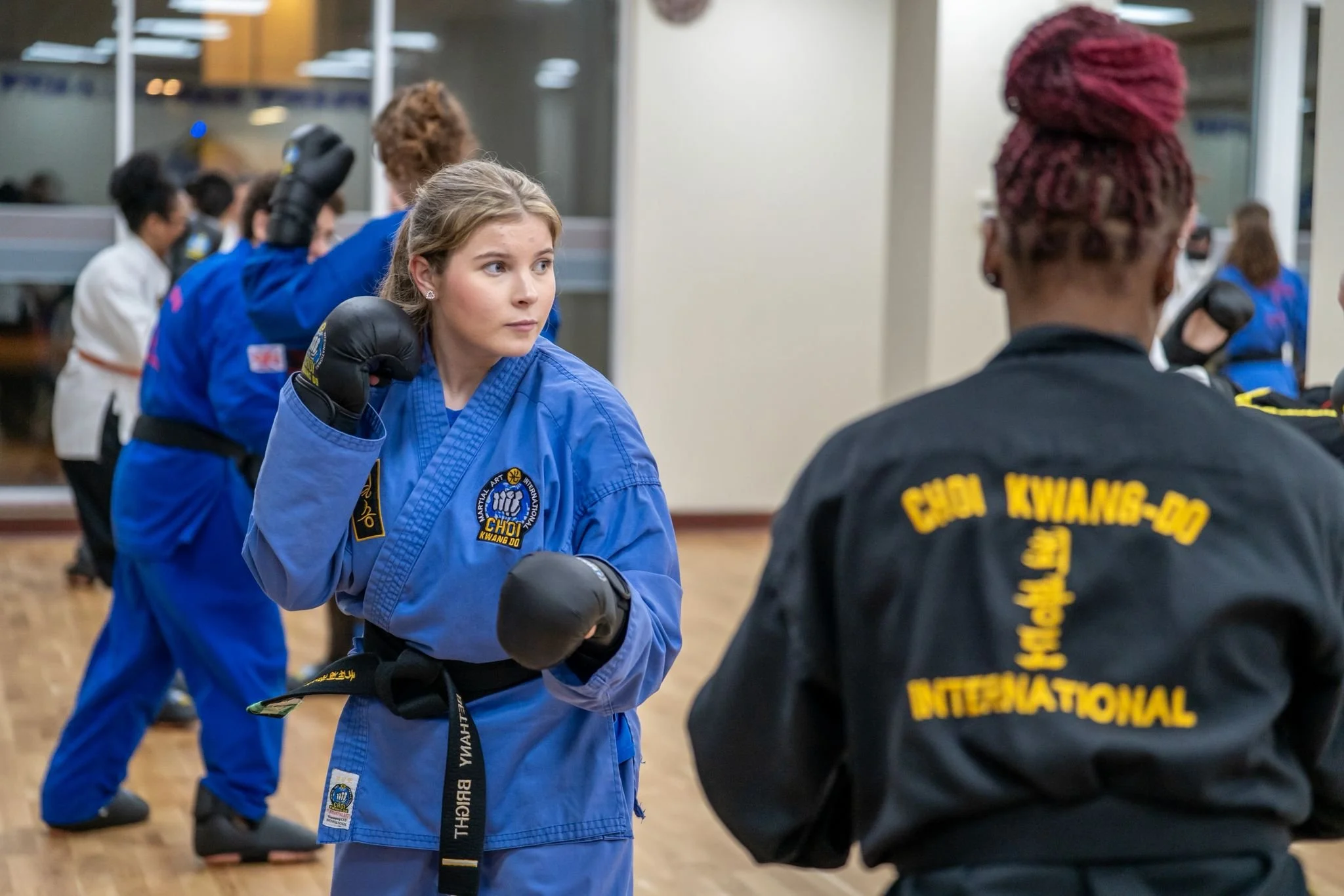 Martial arts students practicing in a training room with wooden floors. One woman in blue uniform is facing another woman in black uniform, both wearing boxing gloves. Other students in martial arts uniforms are visible in the background.