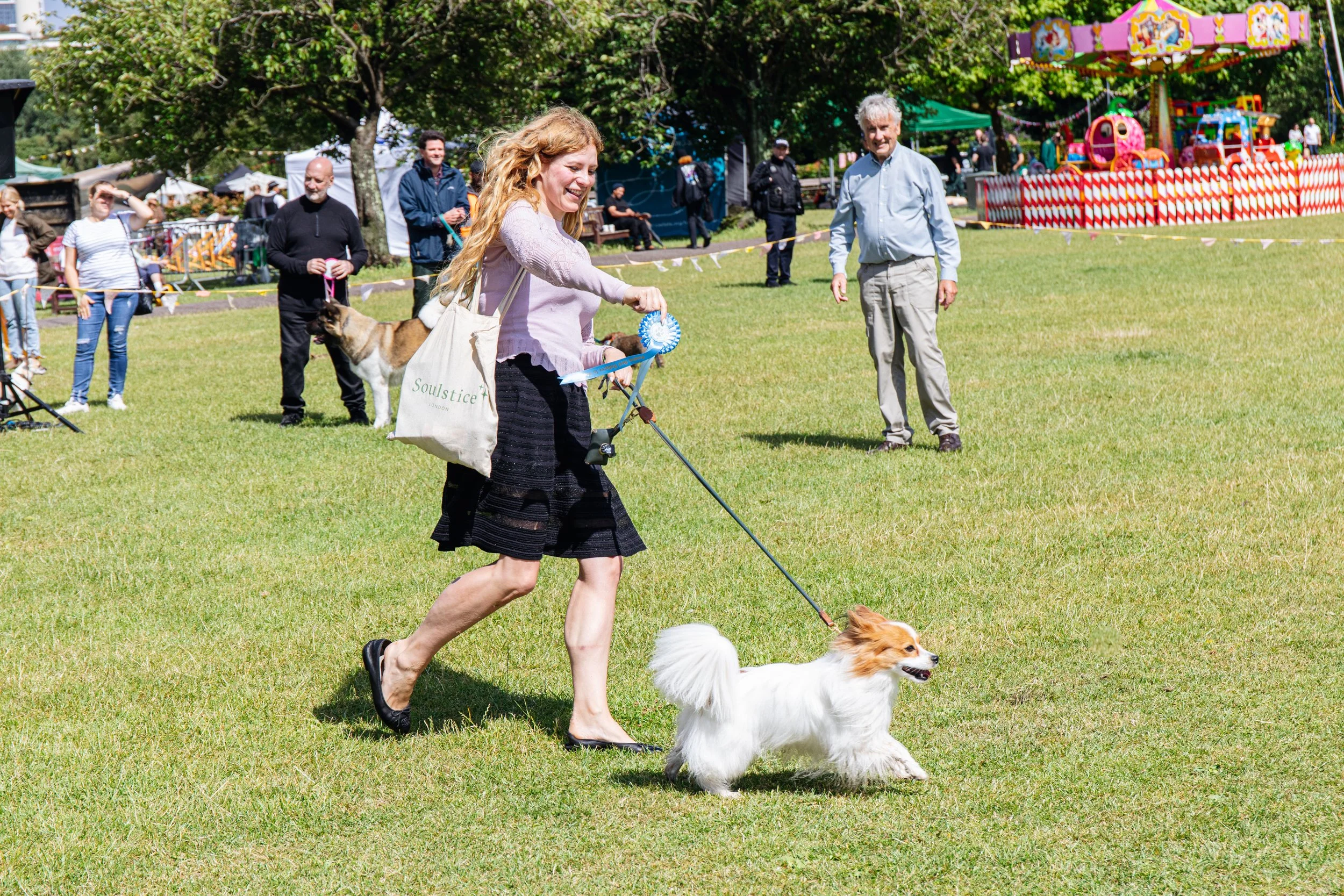 A woman running with a dog on a leash in a park during a sunny day. There is a crowd of people, amusement rides, and trees in the background at a festival or fair.