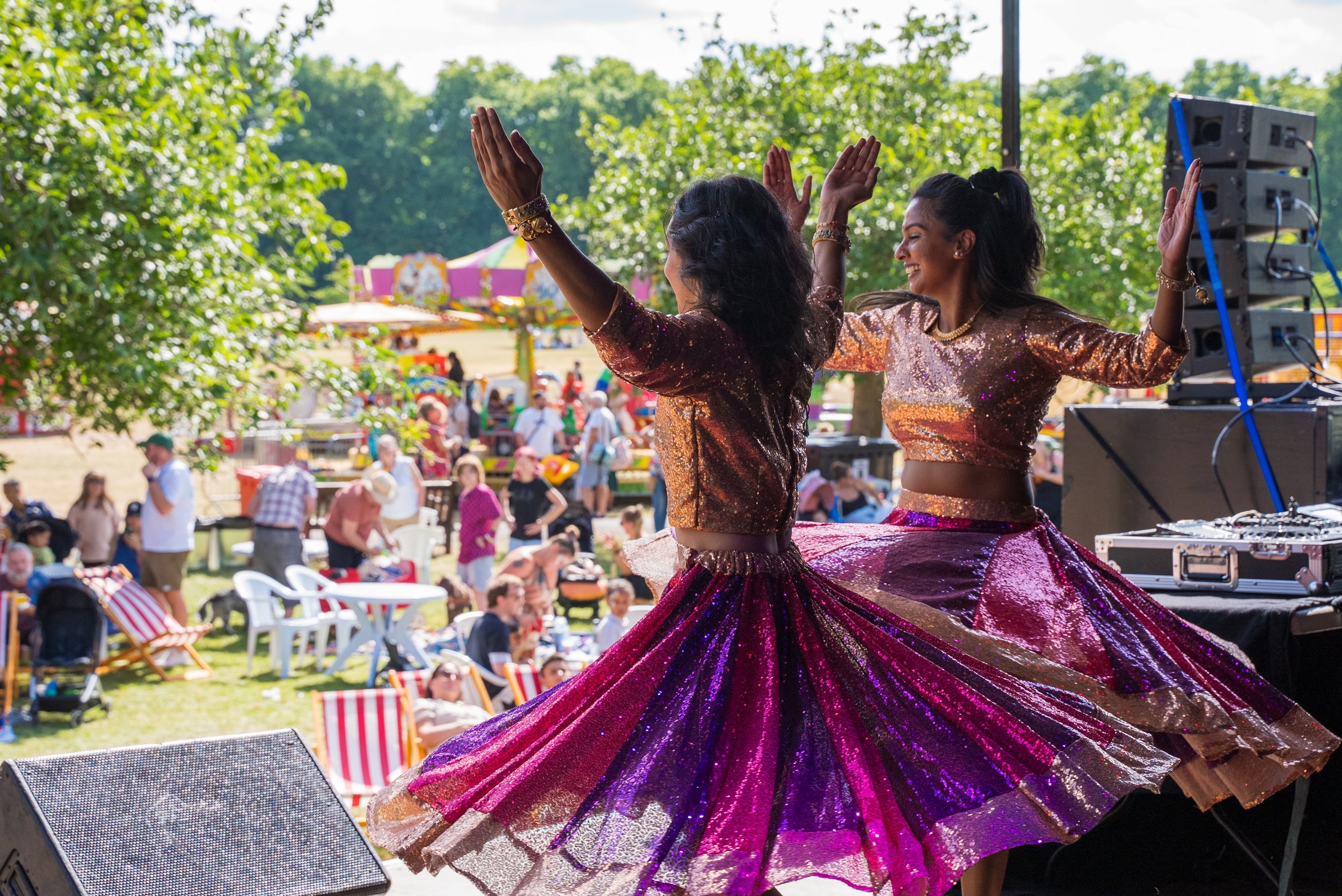 Two women dancing in shiny, colorful dresses at an outdoor event with people, trees, and amusement rides in the background.