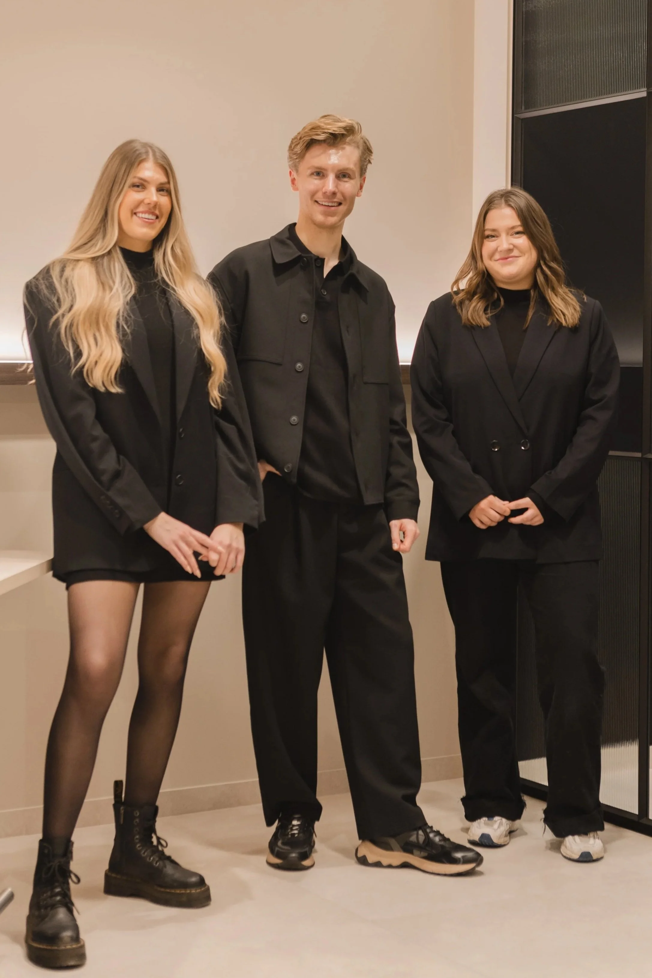 Drietal mensen in zwart gekleed, staand binnen, achtergrond met lichte muur en een kast.