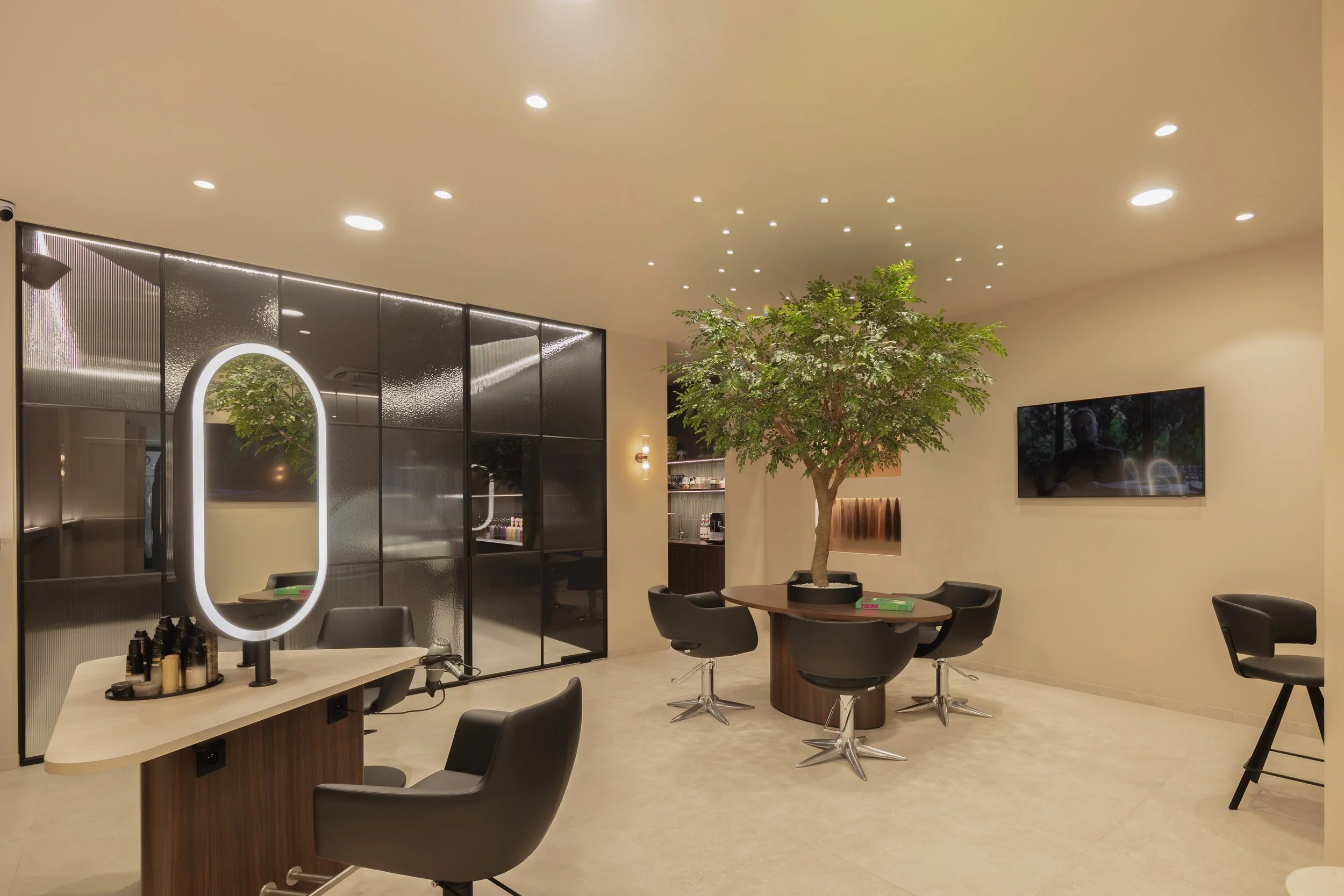 Interieur van een modern kapsalon met een grote boom in het midden, een grote spiegel met verlichting, zwarte stoelen en een tv aan de muur.