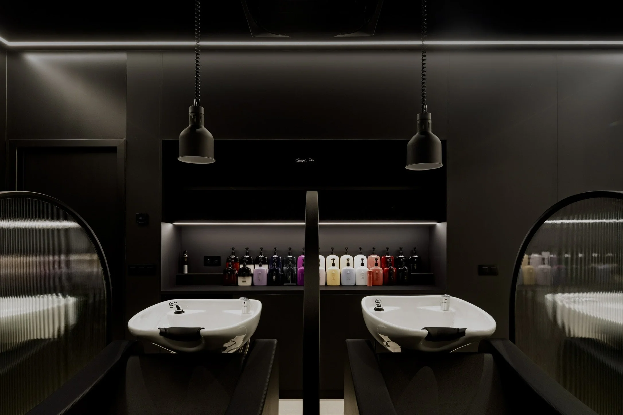Twee wastafels met zwarte beugels, spiegelwanden aan beide zijden, en een schap met diverse gekleurde shampooflessen in een moderne, sfeervolle kapsalon of badkamer.