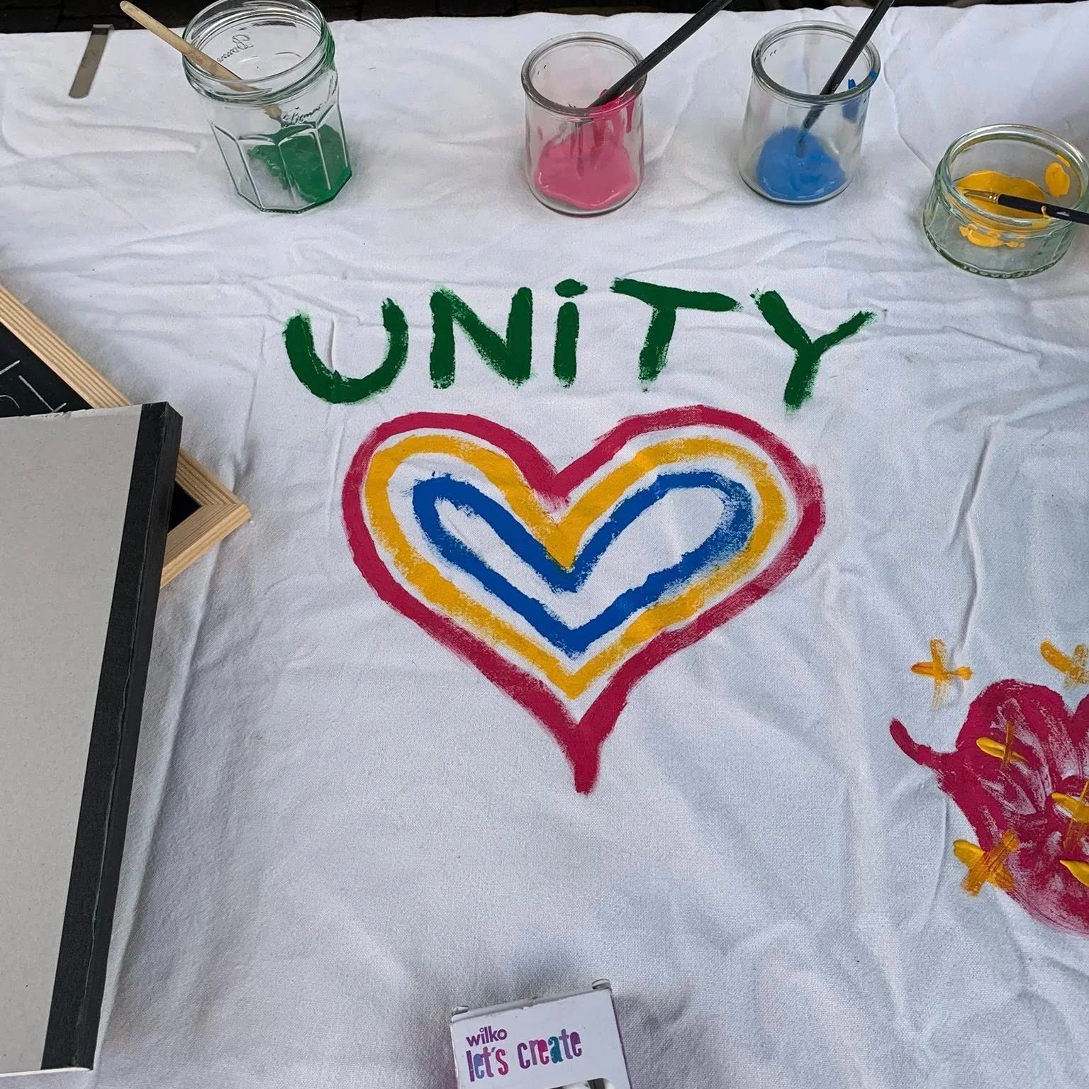 Colorful sign that reads 'UNITY' with a rainbow heart illustration painted below, displayed on a white fabric at an art station with paint jars and brushes.