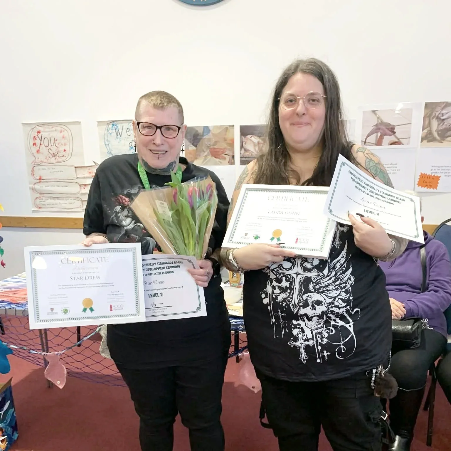 Two women at an award ceremony holding certificates and flowers. The woman on the left has short hair, glasses, and is wearing a dark shirt with a graphic design, while the woman on the right has long hair, glasses, and is wearing a black shirt with white skull print.