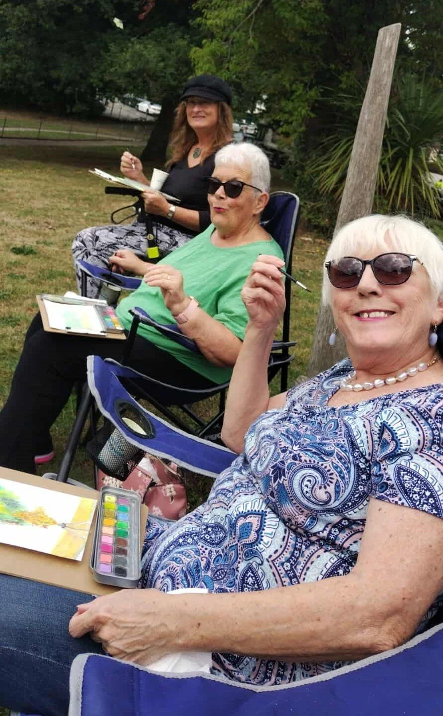 Three elderly women sitting outdoors on camping chairs, smiling and posing for the camera, with art supplies and a painting tablet in front of them, surrounded by trees and grass.