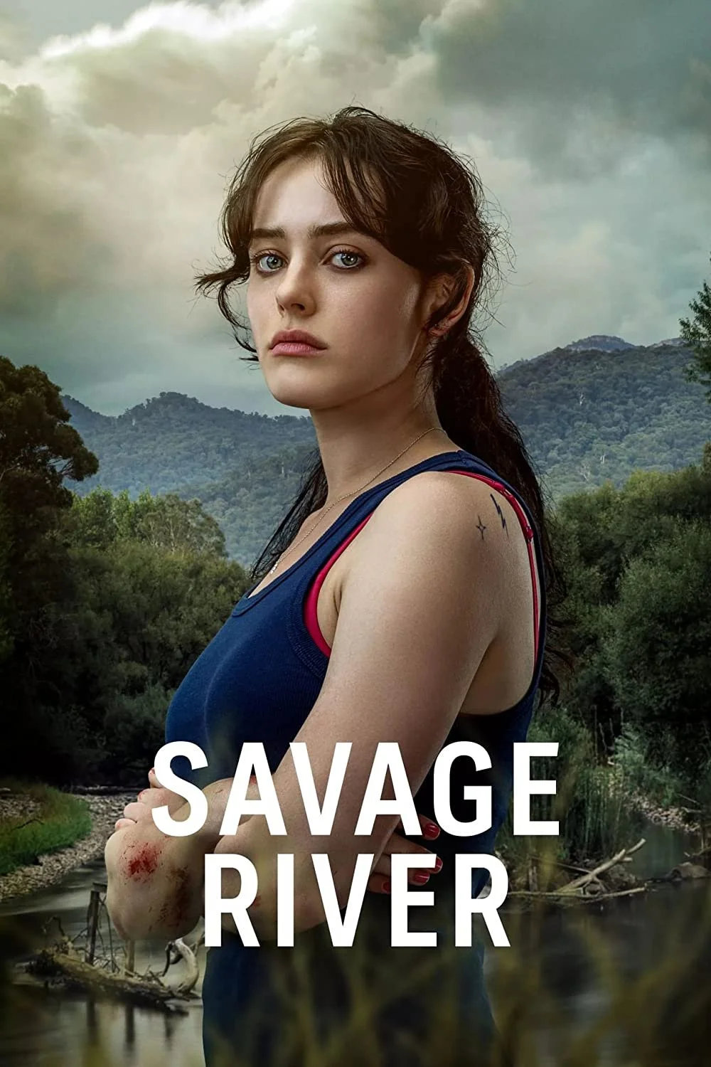 Close-up of a young woman with dark hair and blue eyes standing in front of a river and mountains, with cloudy sky in the background. She has a tattoo on her shoulder and is wearing a blue tank top with red accents. The title 'Savage River' is overla