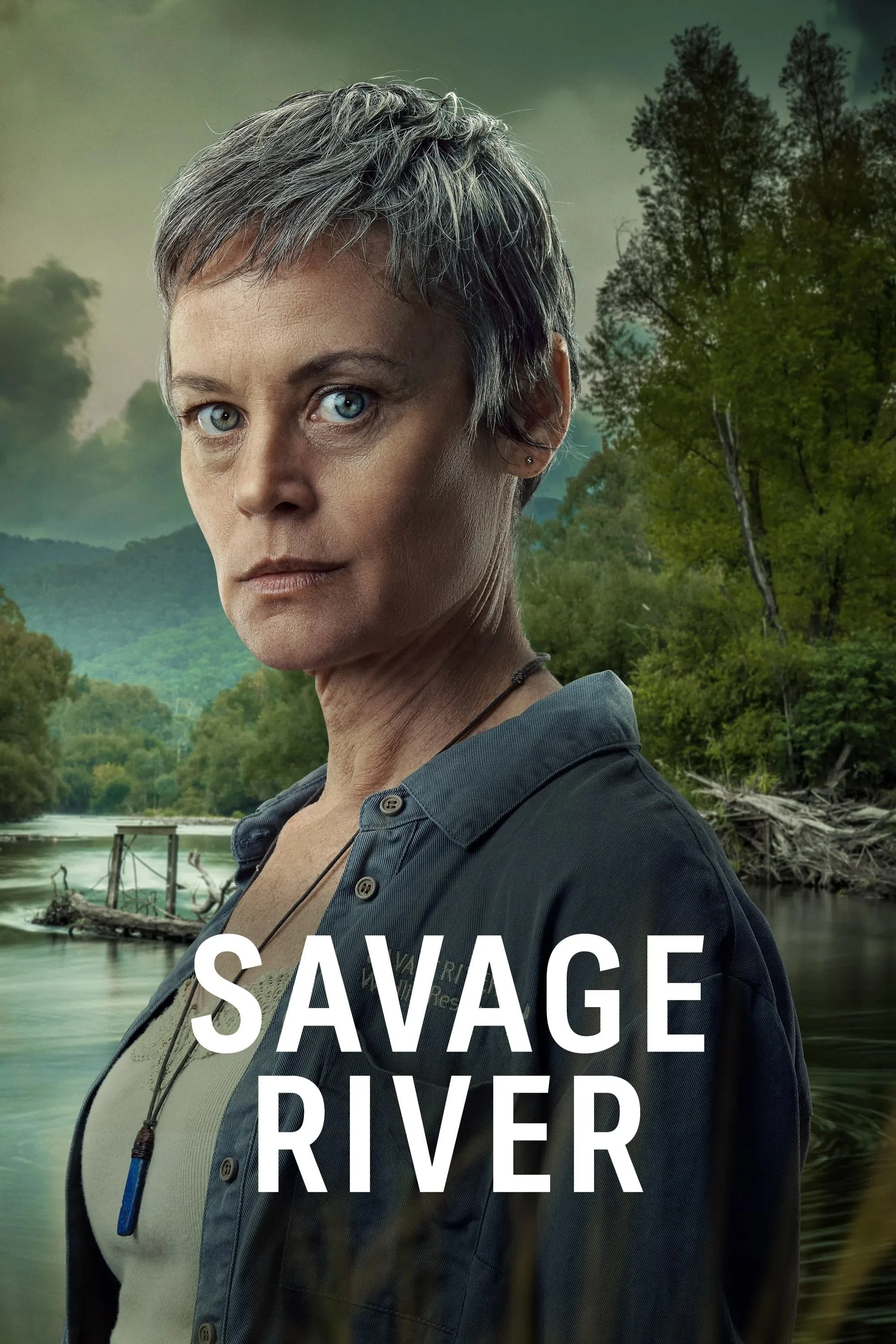 Close-up of an older woman with short gray hair and piercing blue eyes, wearing a dark button-up shirt, standing outdoors with a river and lush green trees in the background, for the TV show 'Savage River'.
