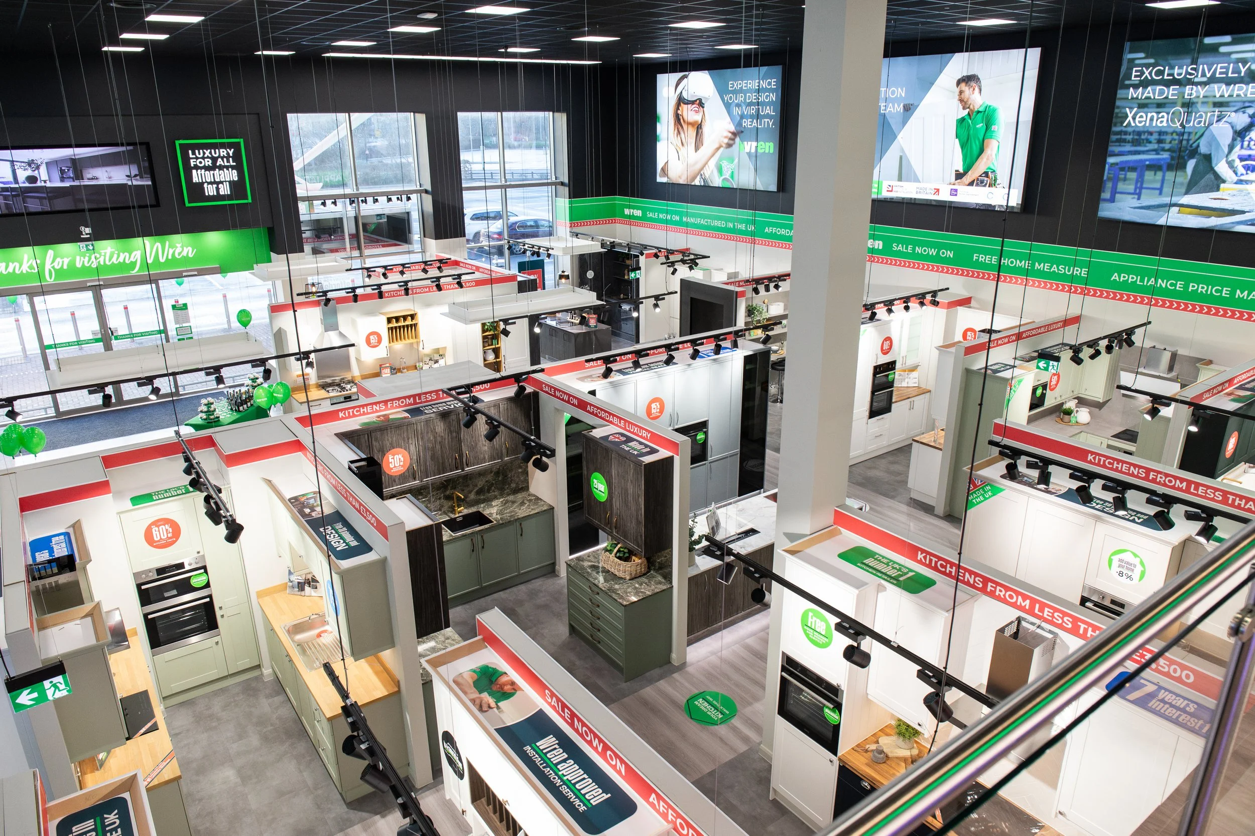 Interior of a kitchen showroom with various kitchen model displays, green signage promoting sales and discounts, overhead lighting, and large windows.