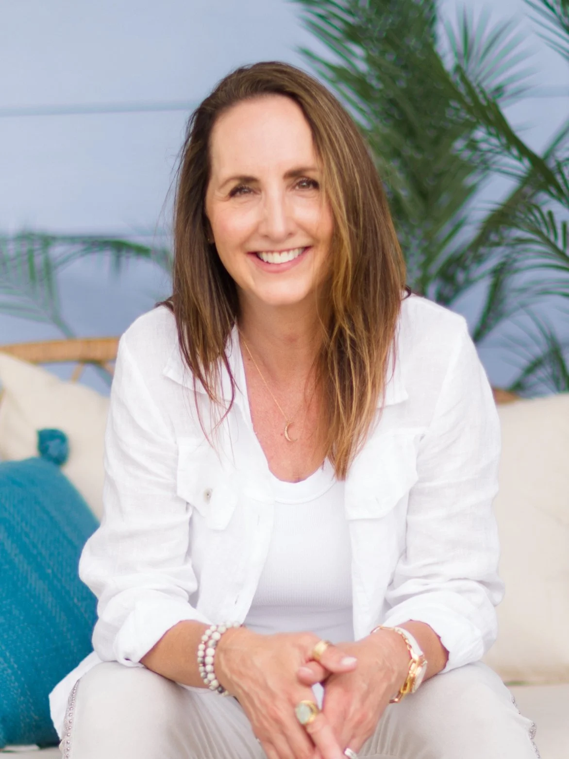 Smiling woman with brown hair sitting indoors near a large green plant, wearing a white jacket, a white top, and jewelry.