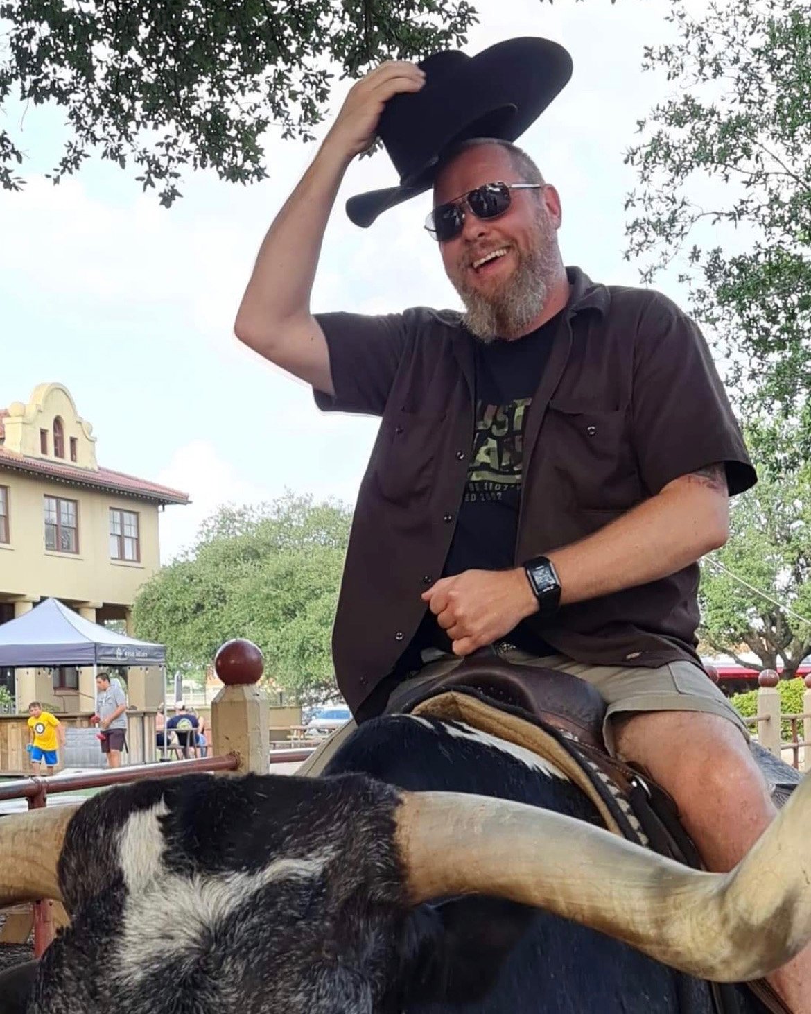 Team member in sunglasses raising a cowboy hat while sitting on a longhorn steer