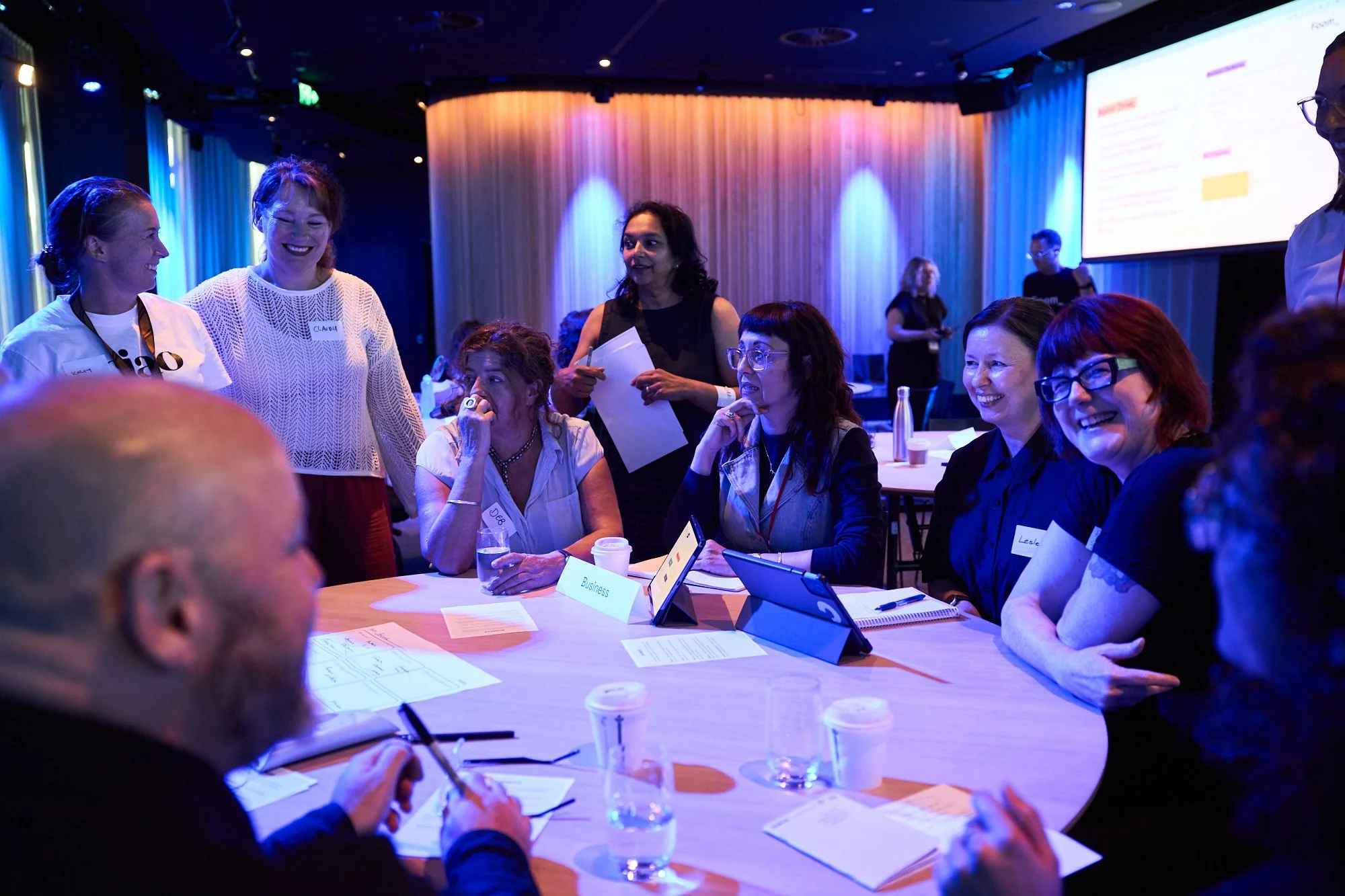 Group of women and men engaged in conversation at a round table during a professional conference or workshop, with conference materials and electronic devices on the table, in a room with colorful lighting and a large projection screen.