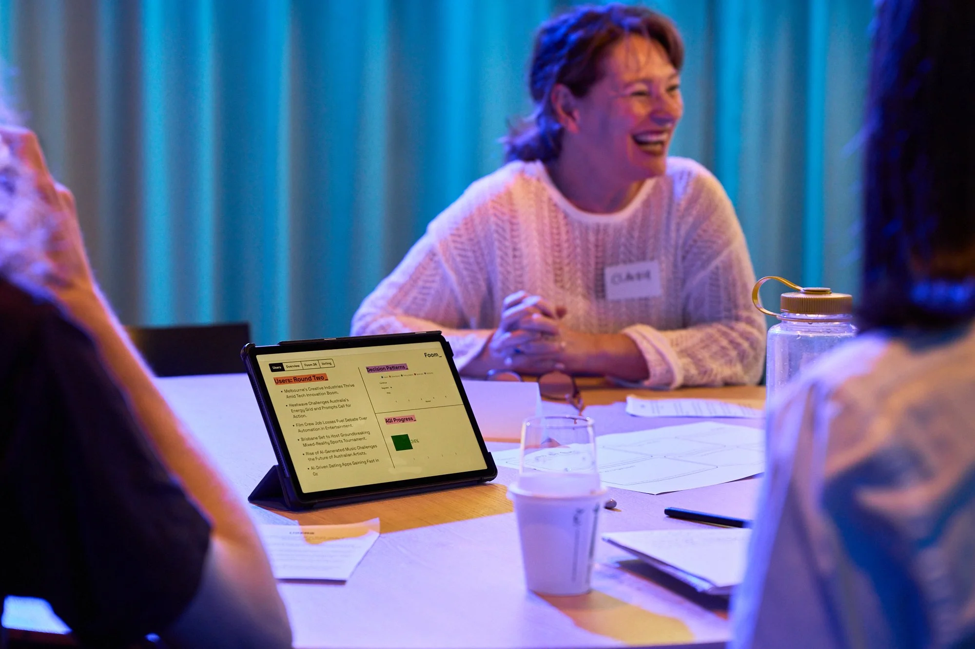 A woman with short brown hair, smiling and laughing, sitting at a table with a group of people in a dimly lit room with blue lighting. She is wearing a light-colored sweater and has a name tag on. There are papers, a tablet, and drinks on the table.