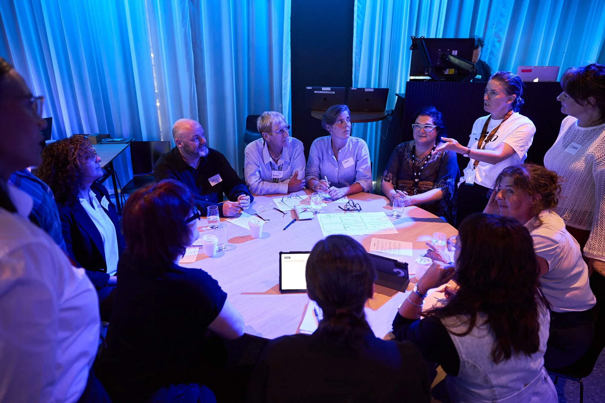A group of people gathered around a round conference table, engaging in discussion. A woman standing on the right side appears to be leading or presenting. The room has blue lighting, with some participants wearing name tags, and various papers, glasses of water, and electronic devices on the table.