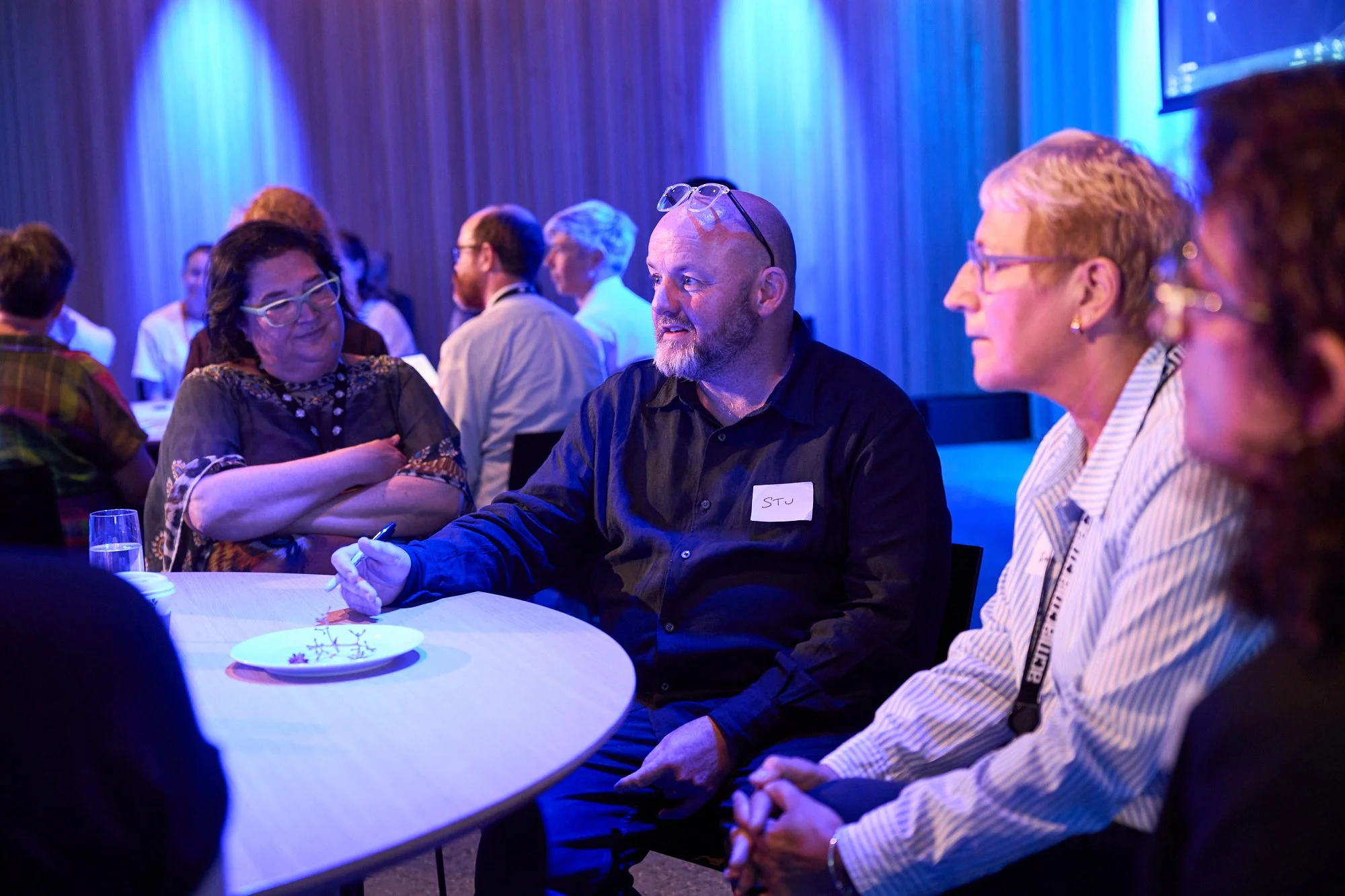 A group of diverse people sitting around a table at an event or conference, engaging in conversation in a room with blue ambient lighting.
