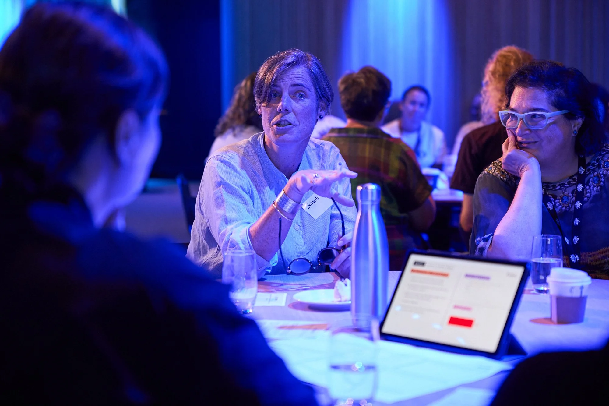 A woman with short with gray hair and a name tag labeled 'Jorie' is talking to someone, sitting at a table with glasses, a water bottle, a laptop, and papers. Other people are in the background, engaging in conversation, in a dimly lit room with blue lighting.