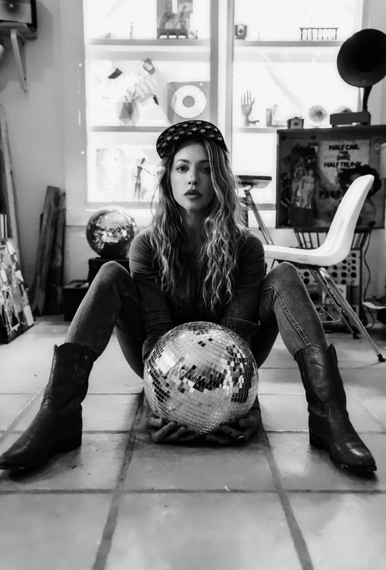 A woman sits on the floor with her legs spread wide, holding a disco ball between her feet. She has long wavy hair, wears a cap, a jacket, jeans, and boots, and gazes into the camera. The background shows a room with large windows, shelves, and various items, including another disco ball.