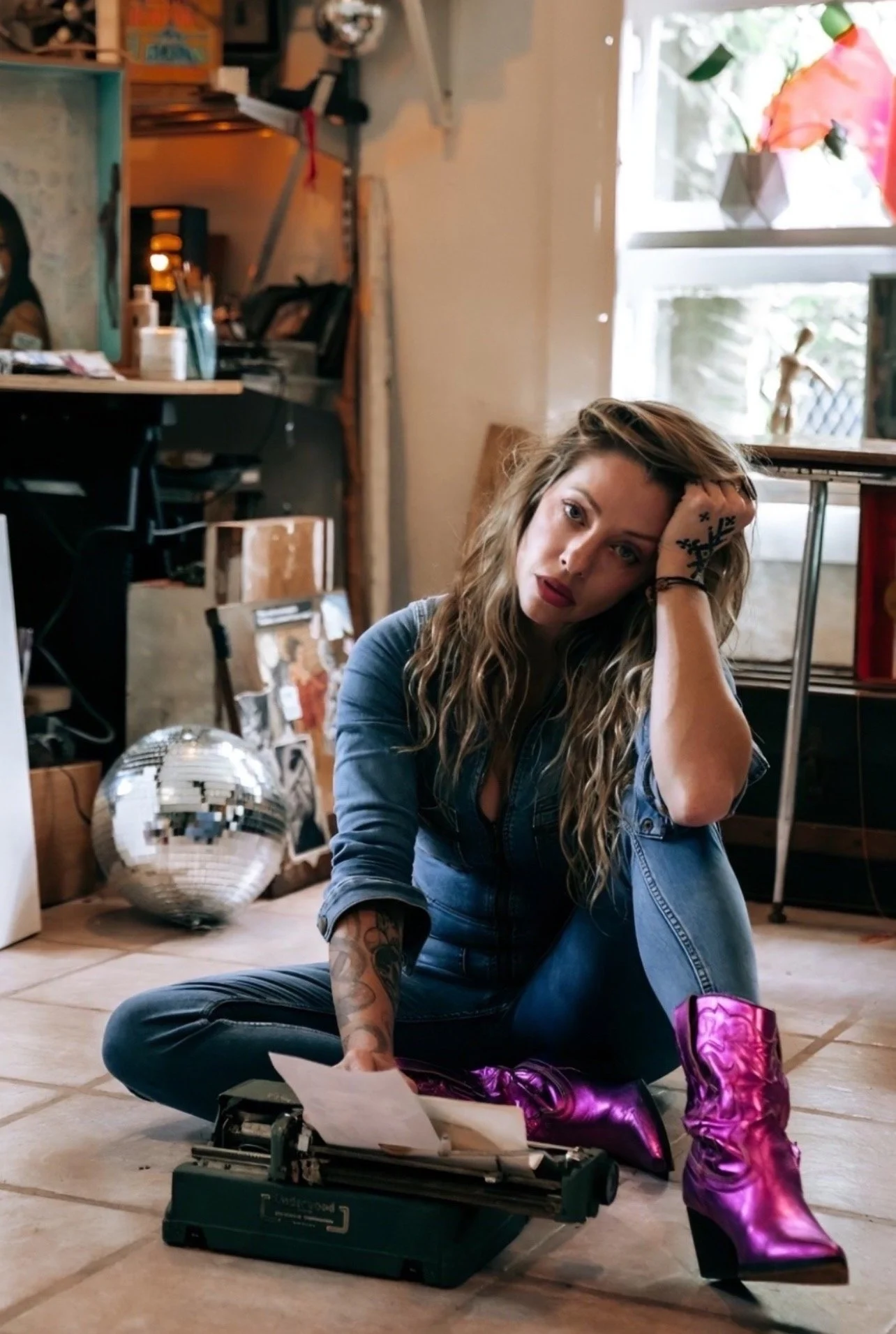 A woman with curly blonde hair and tattoos, sitting on the floor in a cluttered room, wearing metallic purple boots and blue denim outfit, with vintage typewriter and disco ball, looking at the camera with her head resting on her hand.