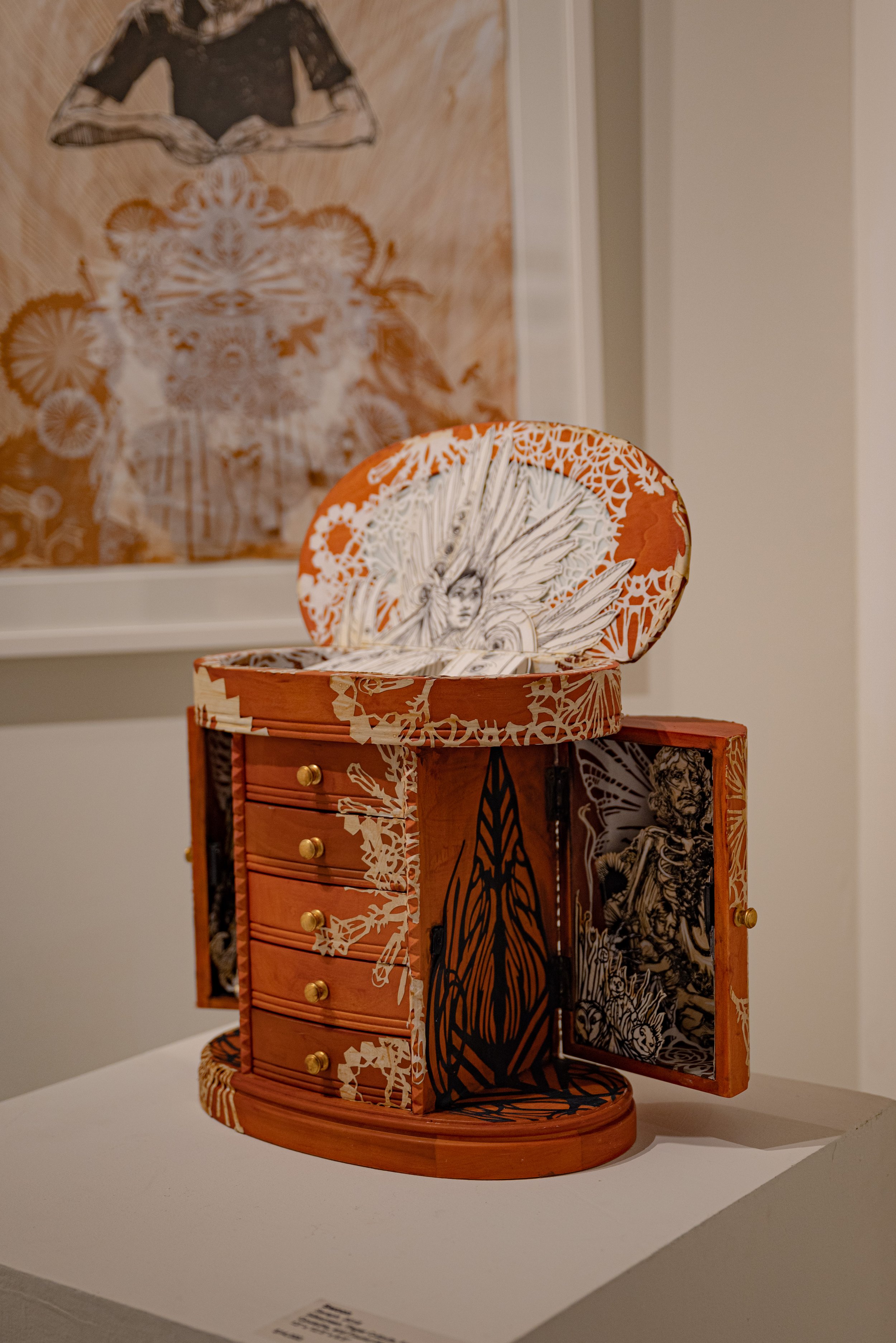 An artistic, miniature wooden jewelry box with intricate drawings and patterns, including a face with wings on the inside of the lid, and a black silhouette of a pointed object on the front. The box has four small drawers with gold knobs and opens to