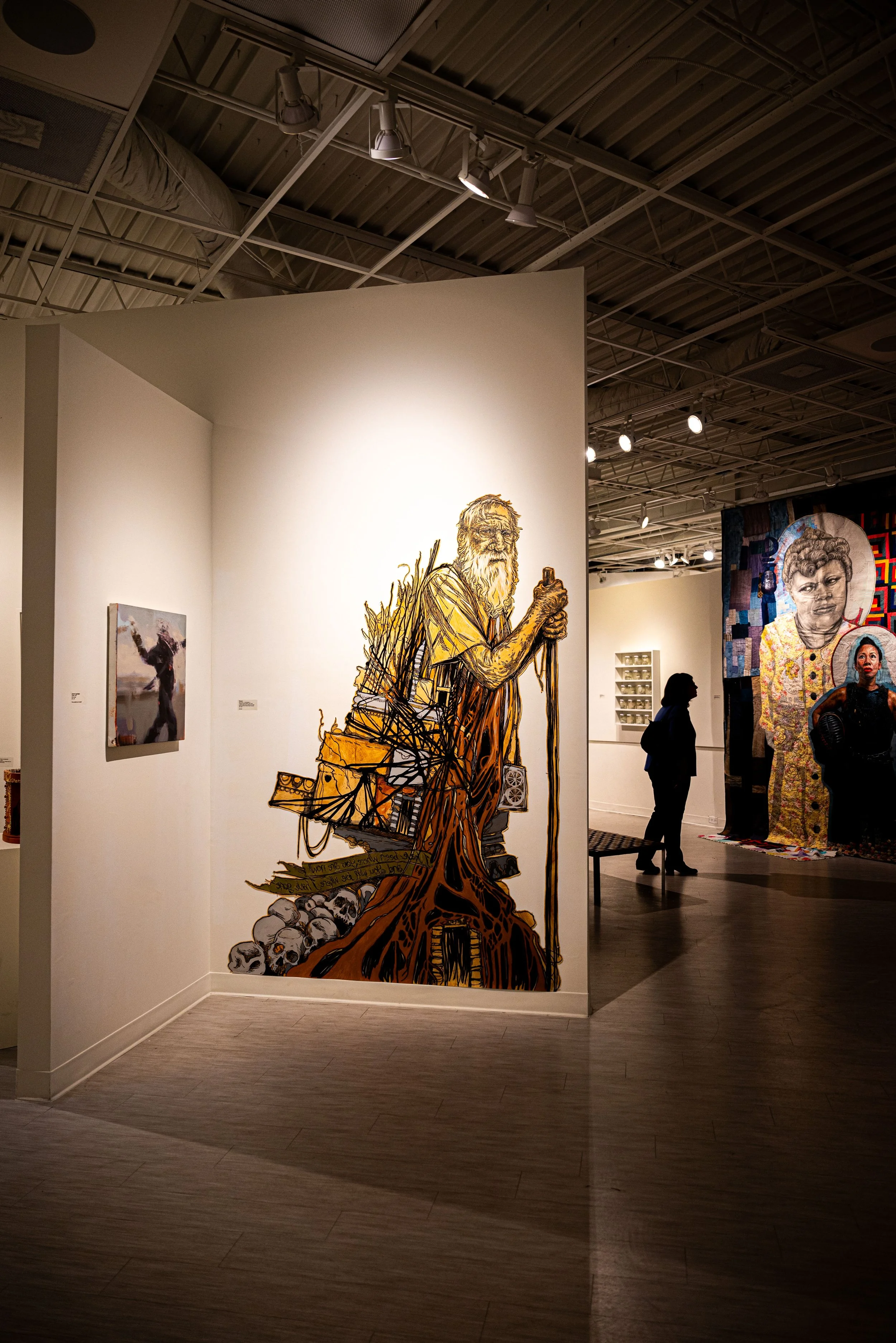 A large artwork of an elderly man with a beard, holding a staff, surrounded by skulls and various objects, displayed on a white wall in an art gallery.