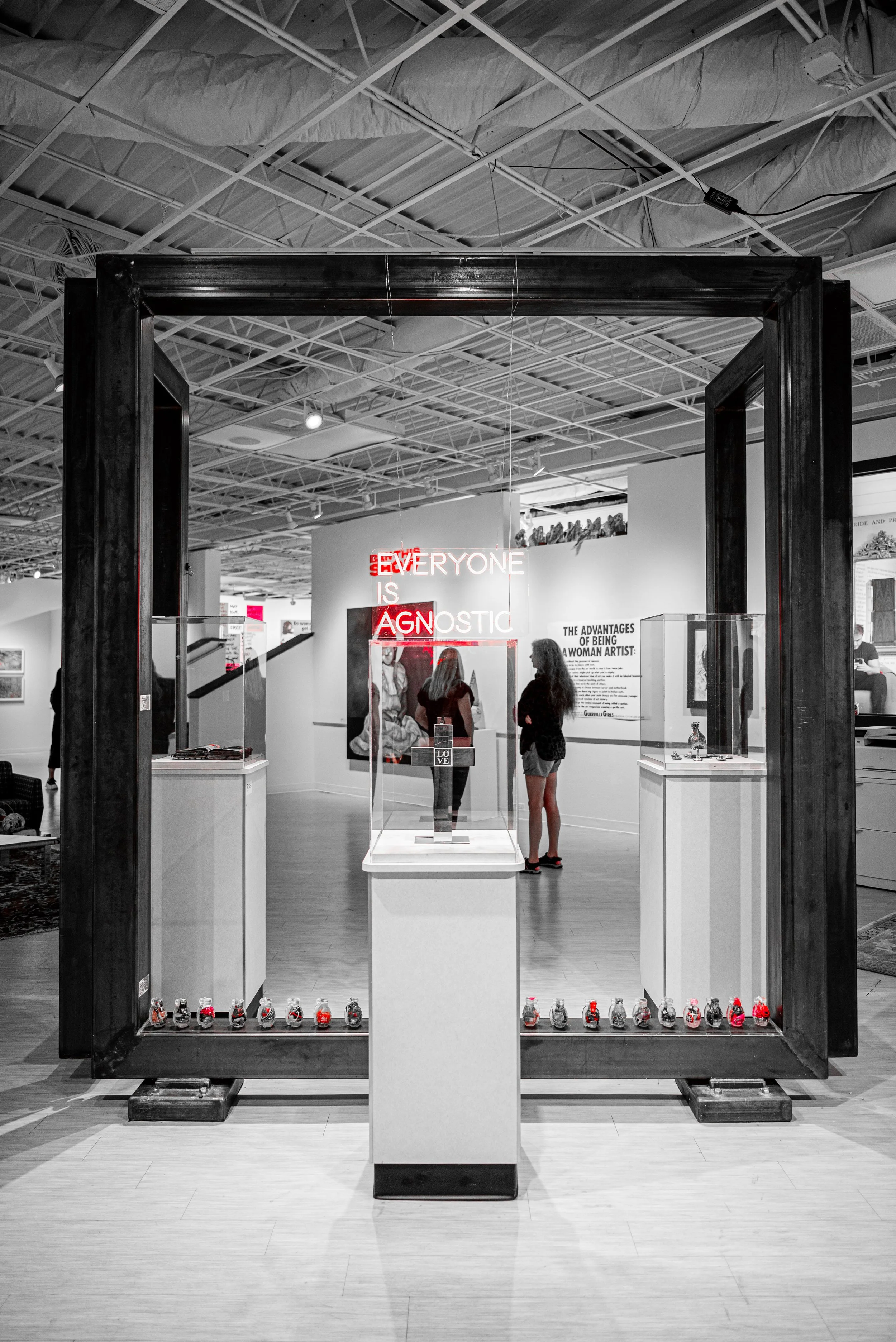 An art exhibit features a neon sign reading 'EVERYONE IS AN OPTIC' in red and white, with women observing artwork behind the glass display case, and small art pieces arranged along the floor and on display pedestals.