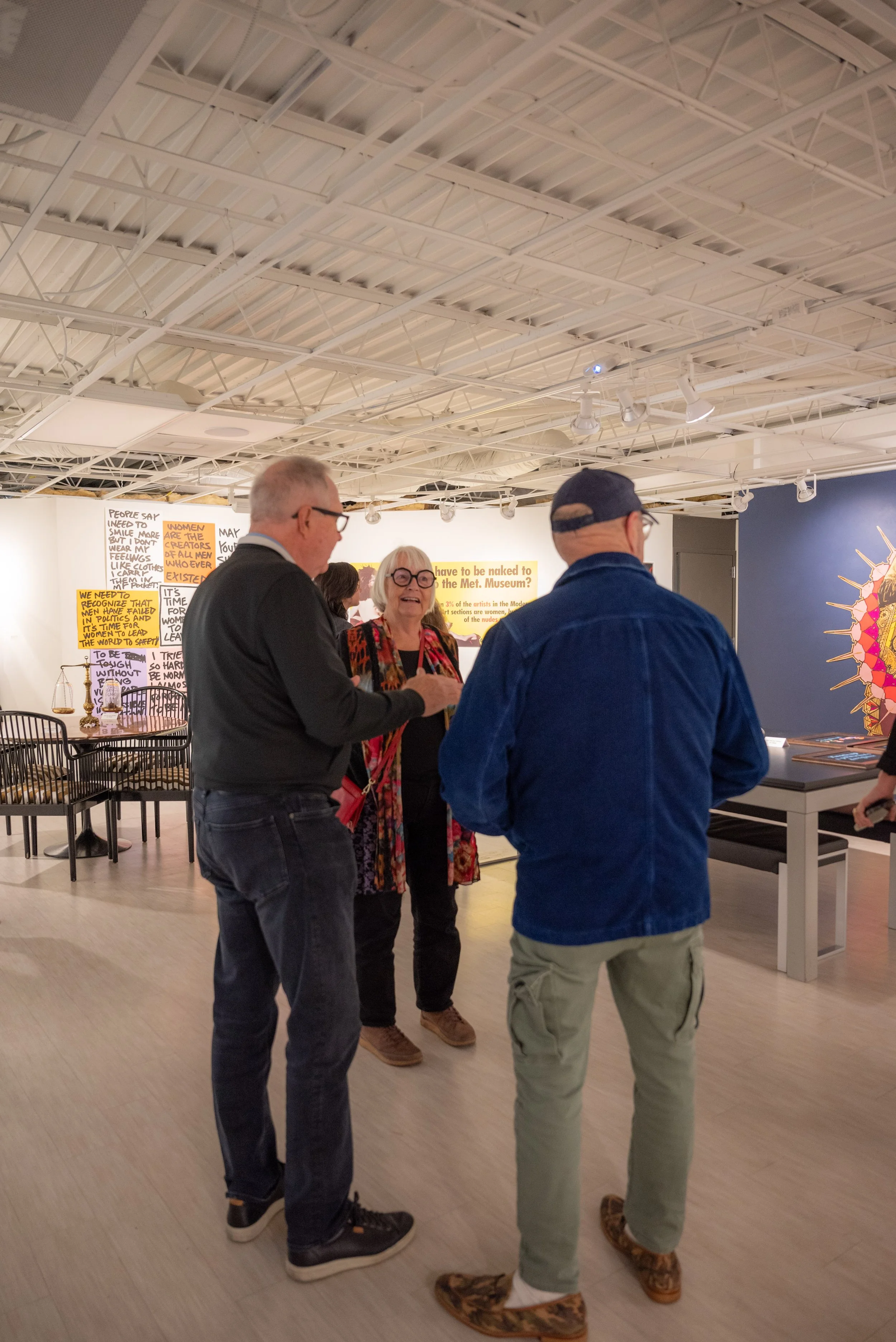 Three people stand and talk inside an art museum with contemporary artwork and yellow text panels on the wall.