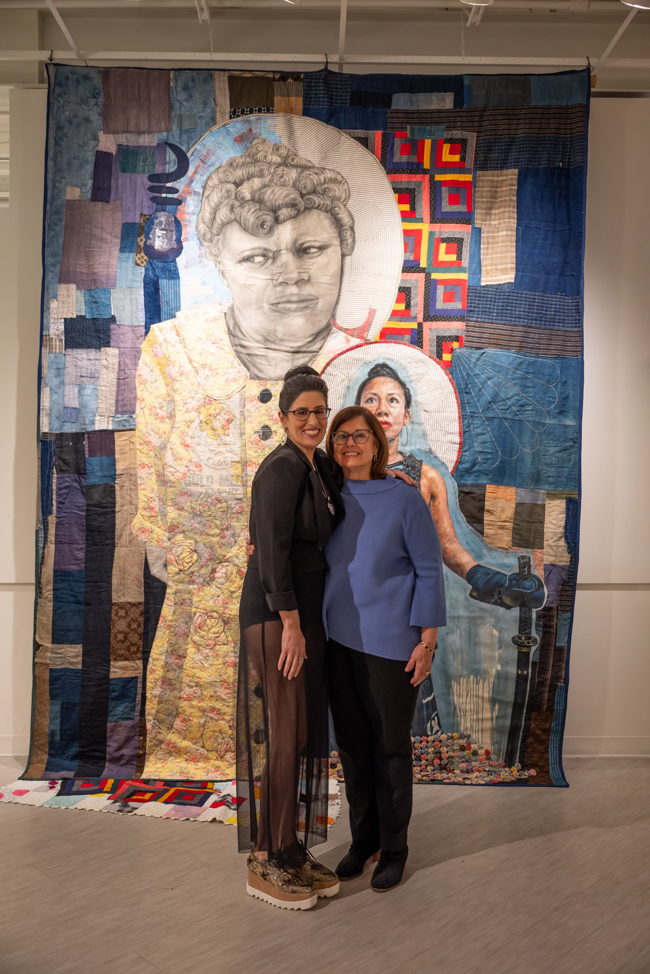 Two women standing close together, smiling, in front of a large quilt portraying two women, one with a large curl hairstyle and a serious expression, and the other with a headscarf and glasses, holding a cane, surrounded by various patchwork fabric p