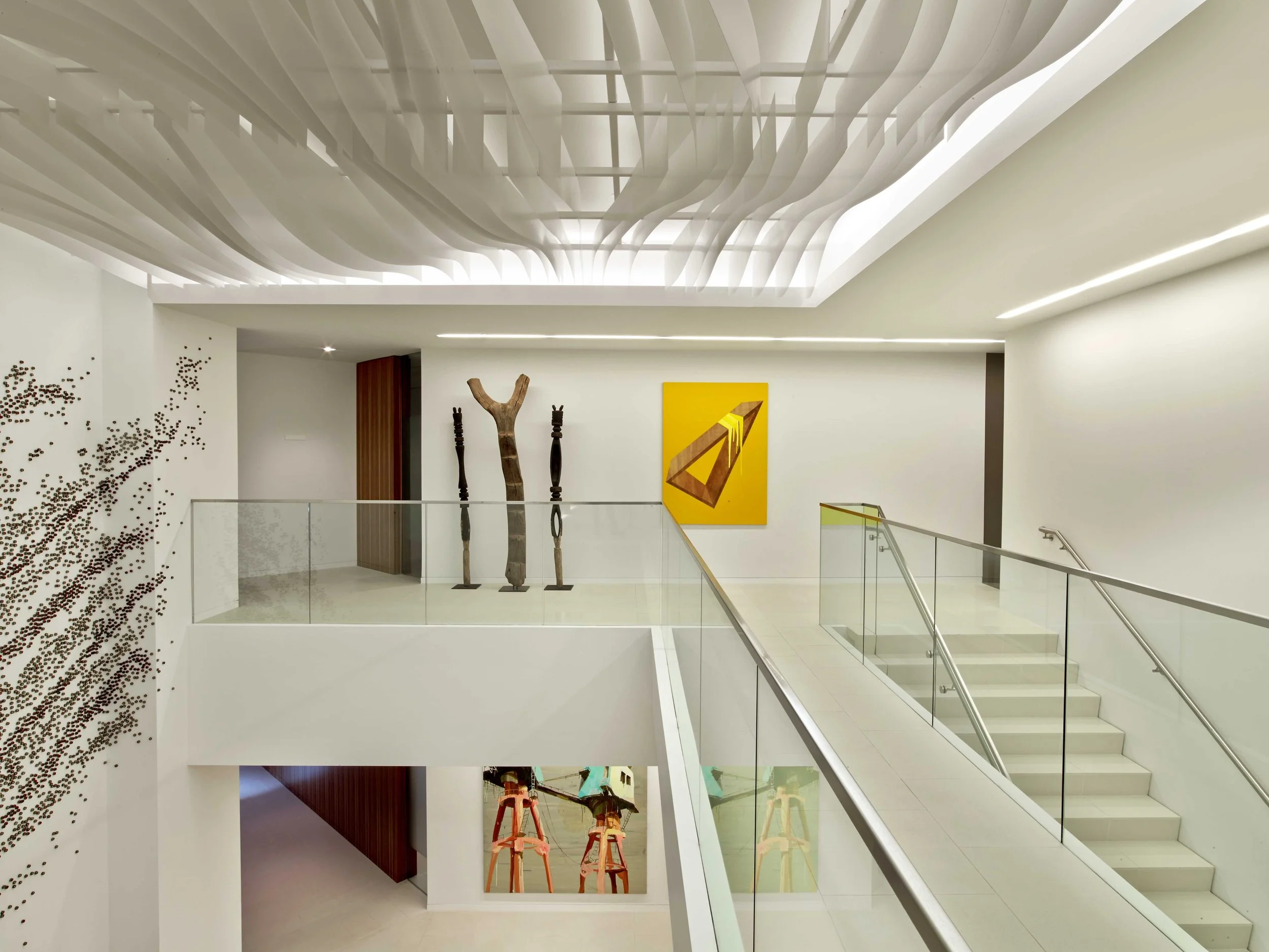 Modern interior of a building with white walls, a staircase with glass railing, and minimalist art pieces including a yellow and brown geometric painting, sculptures resembling tall, slender wooden figures, and contemporary decor elements.