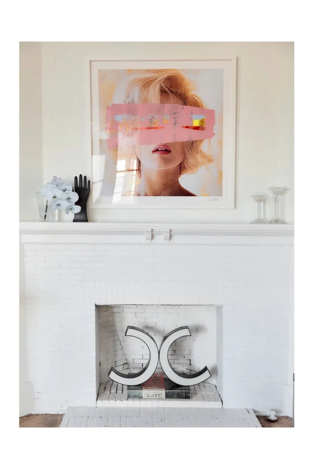 Indoor minimalist decor featuring a white fireplace with abstract sculpture, a black hand-shaped object, a bunch of white flowers, and framed artwork of a woman's portrait with pink painted obscuring her eyes.