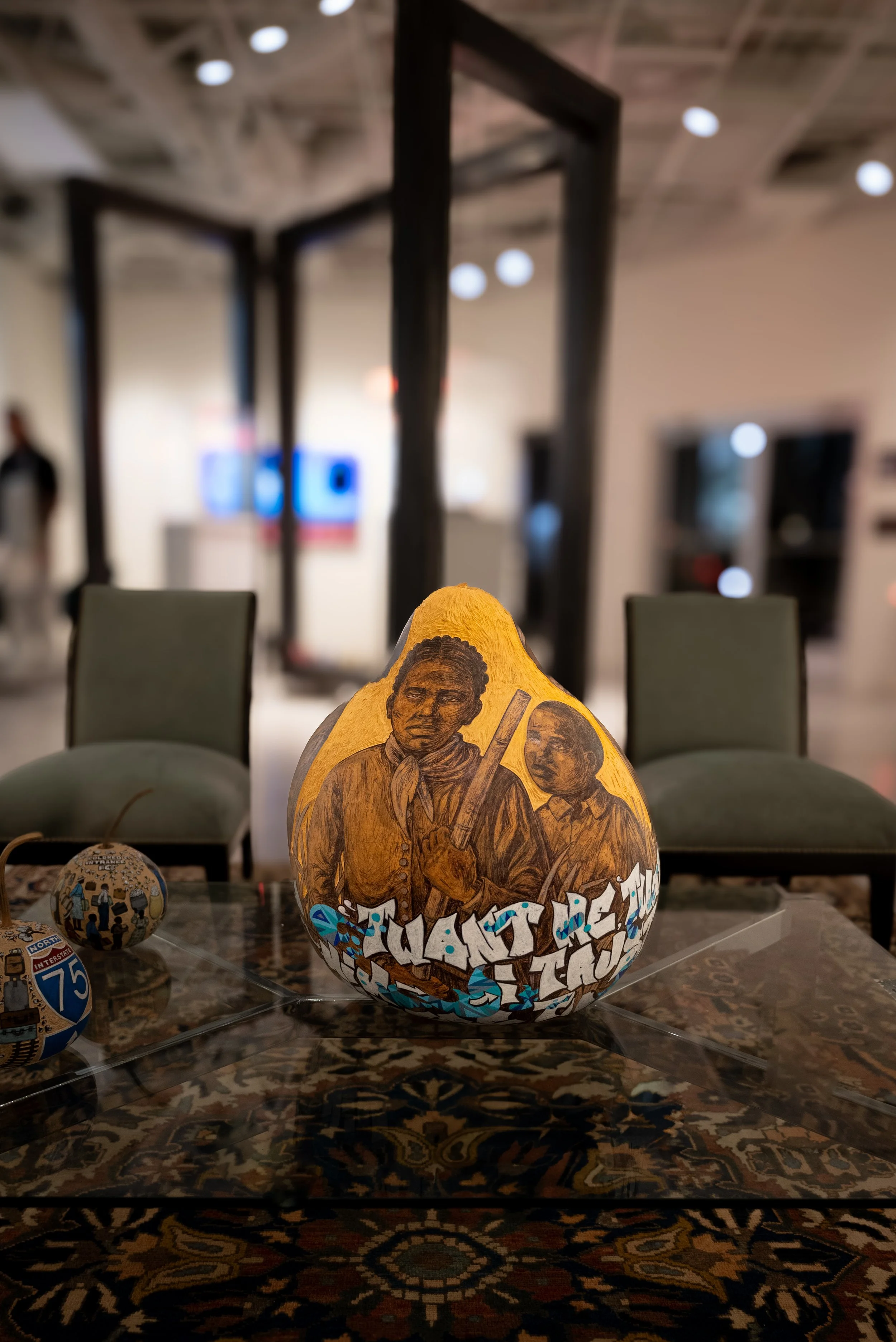 A painted gourd with historical figures and graffiti-style text, displayed on a table in an indoor gallery or museum setting.
