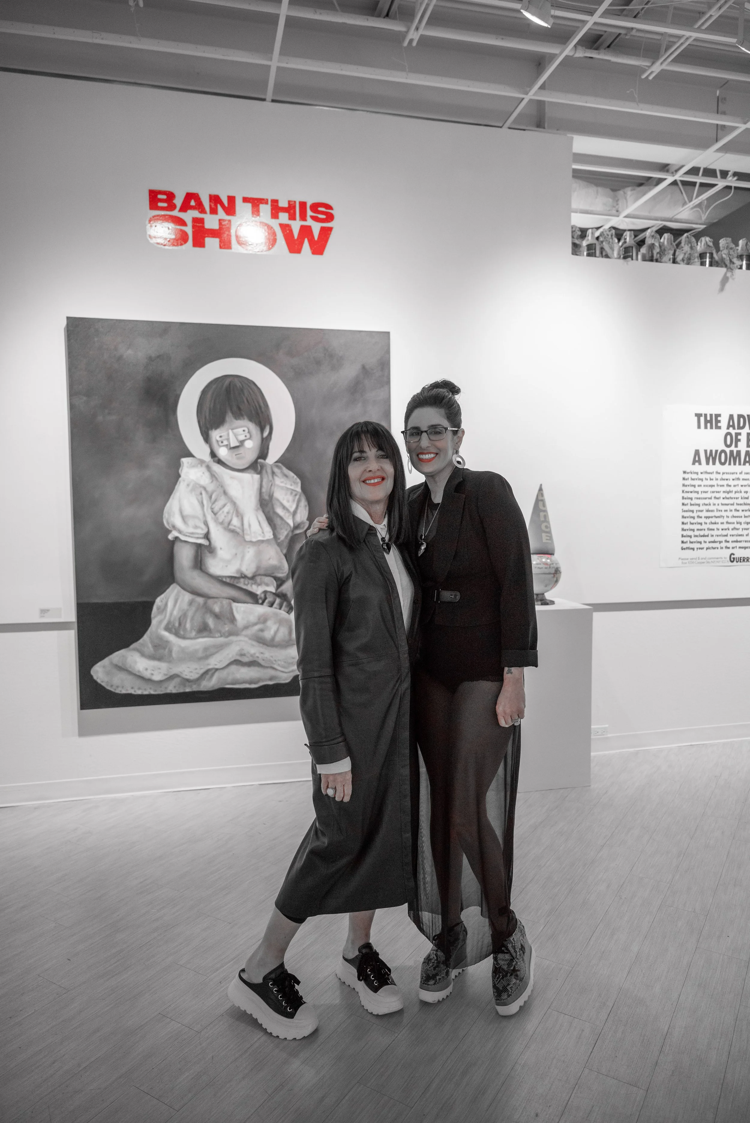 Two women standing together in an art gallery, smiling. Behind them is a painting of a girl with a bob haircut wearing glasses. A sign above reads "BAN THIS SHOW."