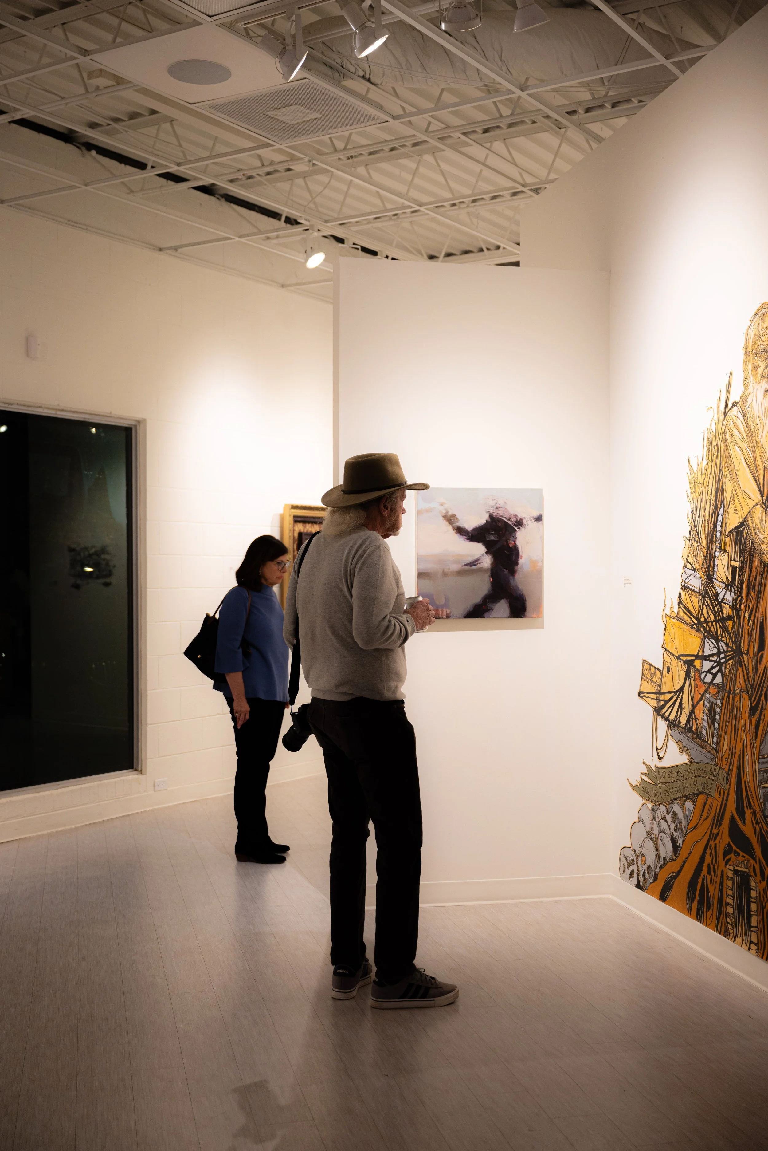 Two women and a man observing artworks at an art gallery. The man has blond hair, a hat, gray sweater, black pants, and sneakers. One woman has dark hair, glasses, a blue top, and is carrying a black bag. The other woman has dark hair, glasses, a dar