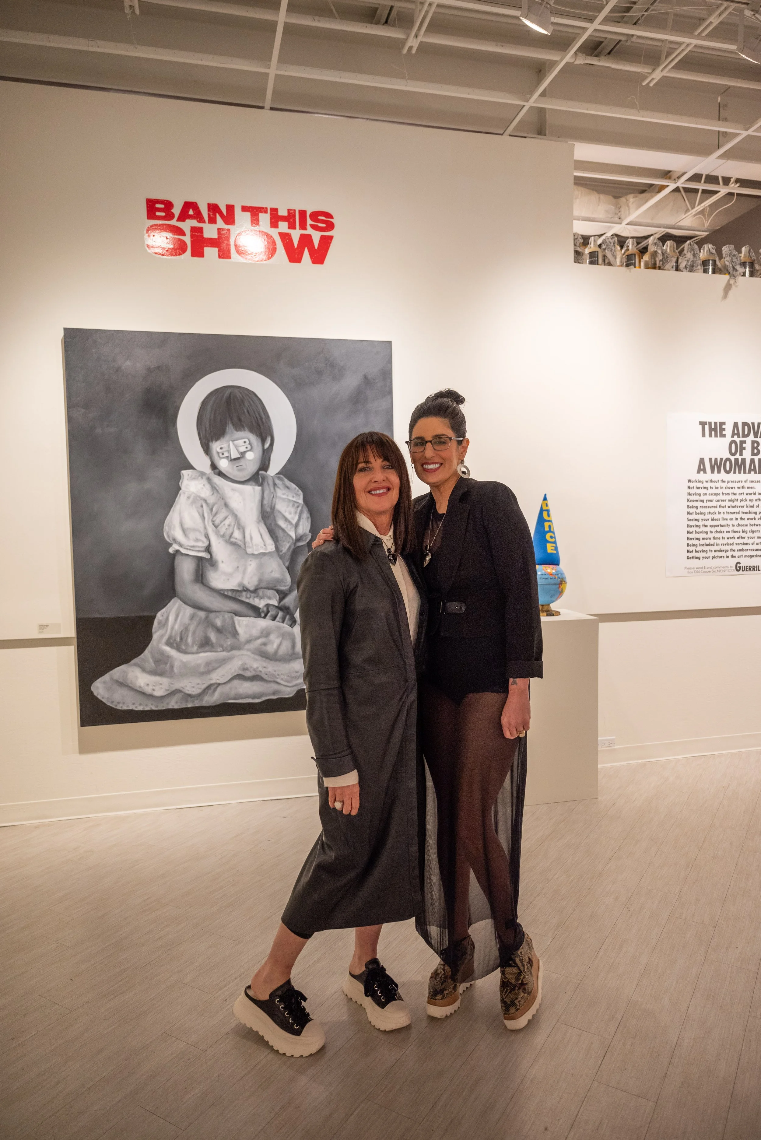 Two women standing together in an art gallery, smiling for the camera. Behind them is a large black and white painting of a girl with a halo and a bandage over her nose, and above the painting is a red sign that says "BANTHISSHOW." To the right, ther