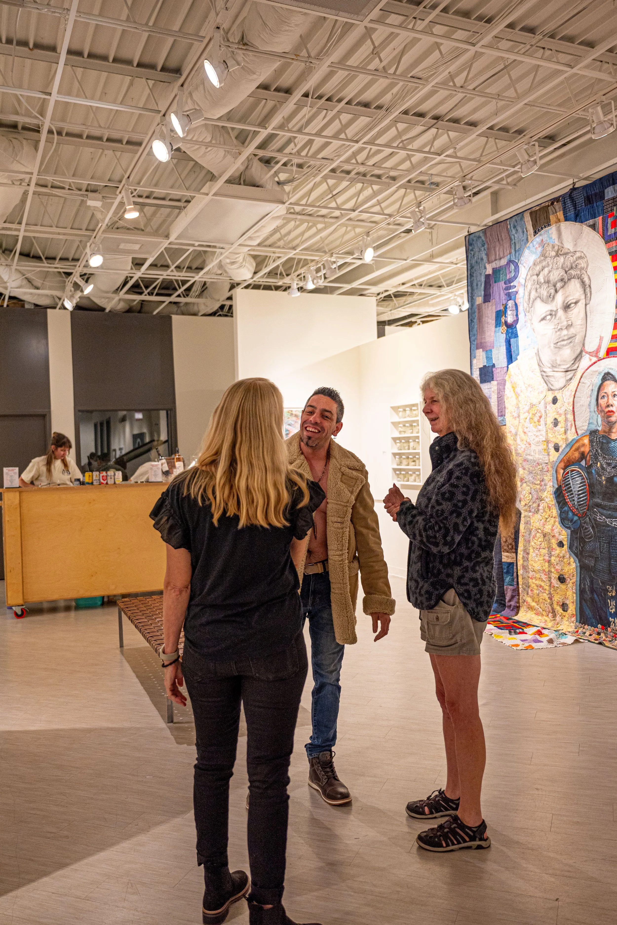 Three people are seen standing in an art gallery, engaged in conversation and smiling, with a large colorful tapestry of a woman's portrait on the wall behind them.