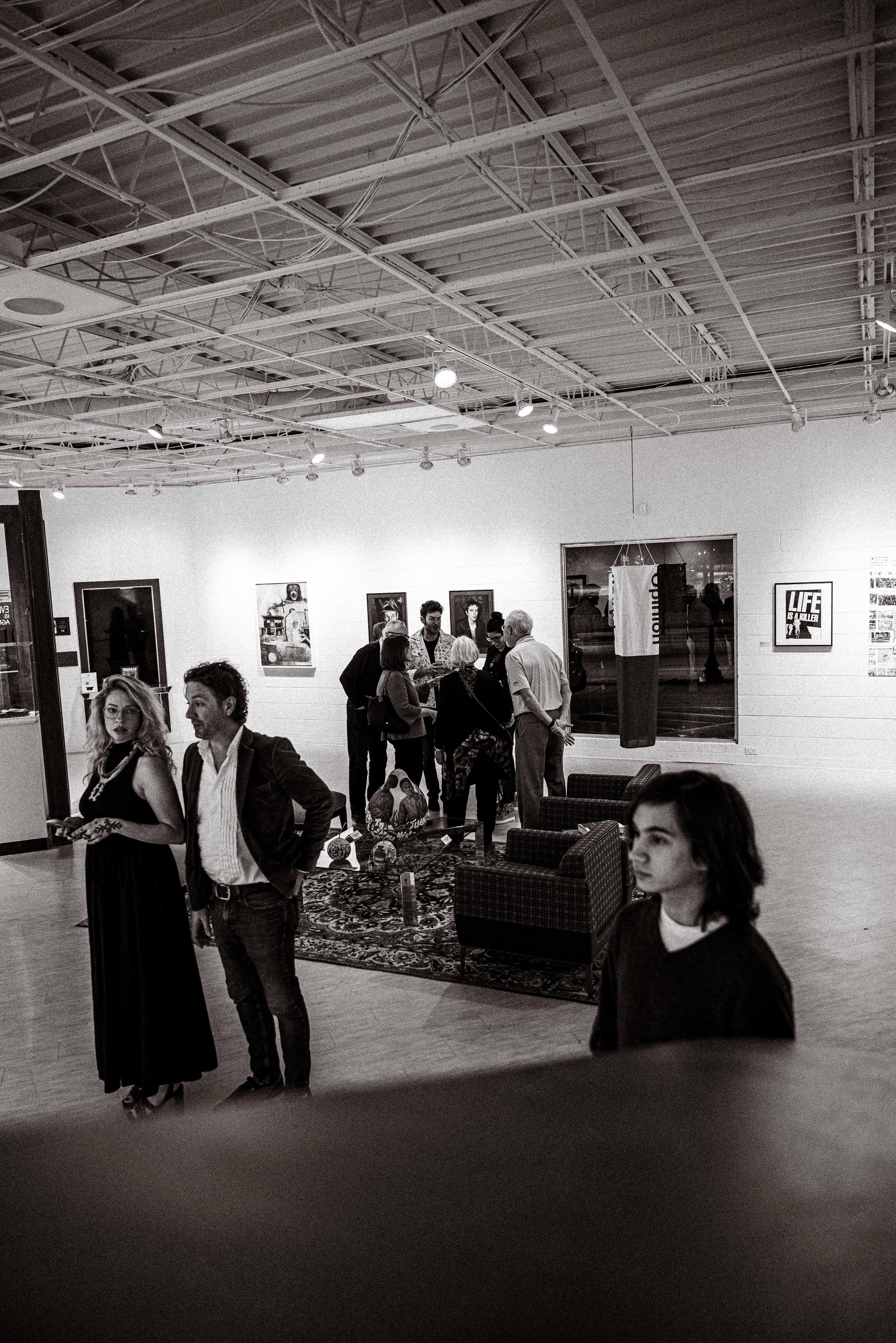 People gathered in an art gallery, viewing paintings and artworks on the walls, with some engaged in conversations.