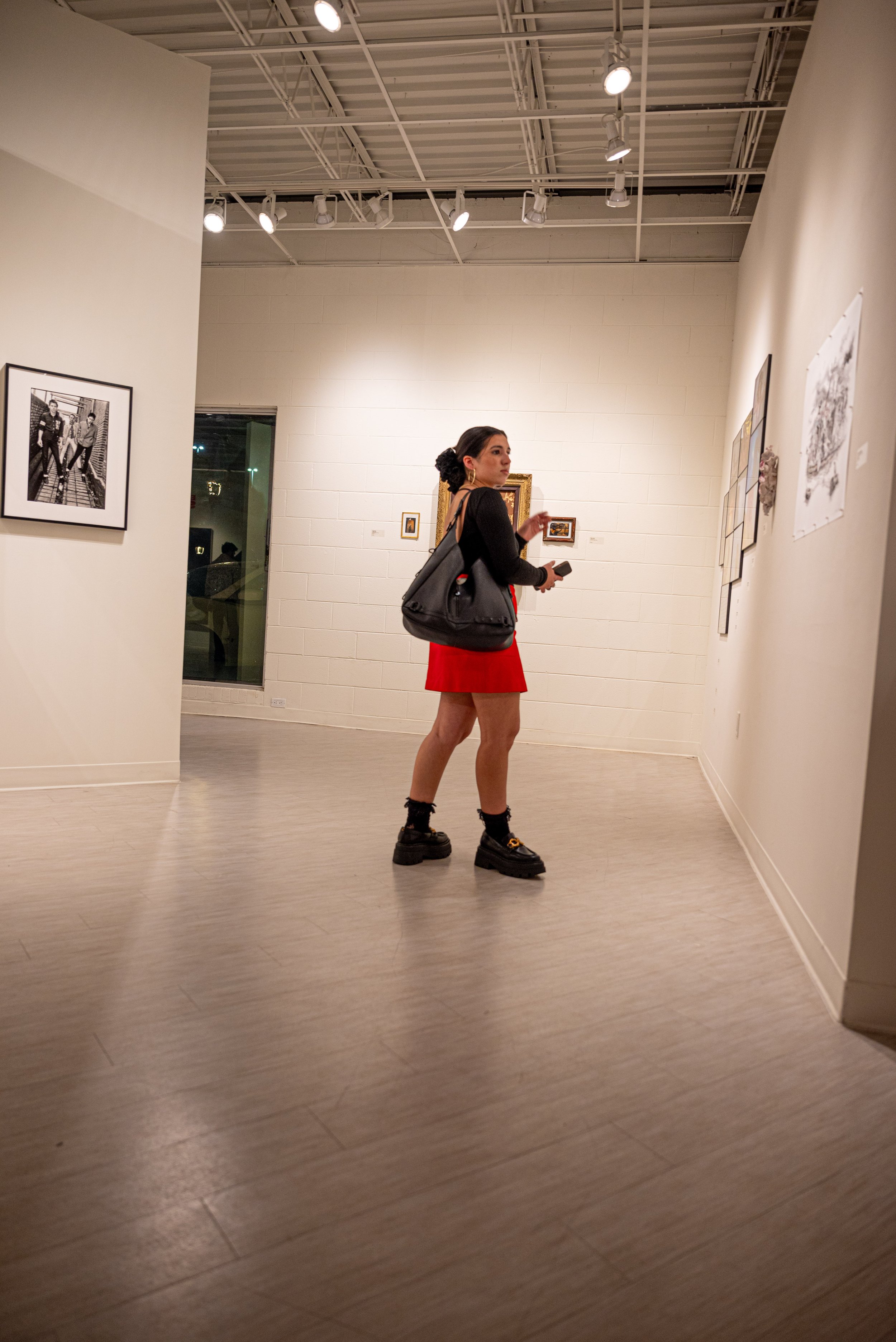 A woman in a black top, red skirt, and chunky black shoes with socks, stands in an art gallery looking at the artwork on the wall. She carries a large black handbag and holds a phone in one hand.