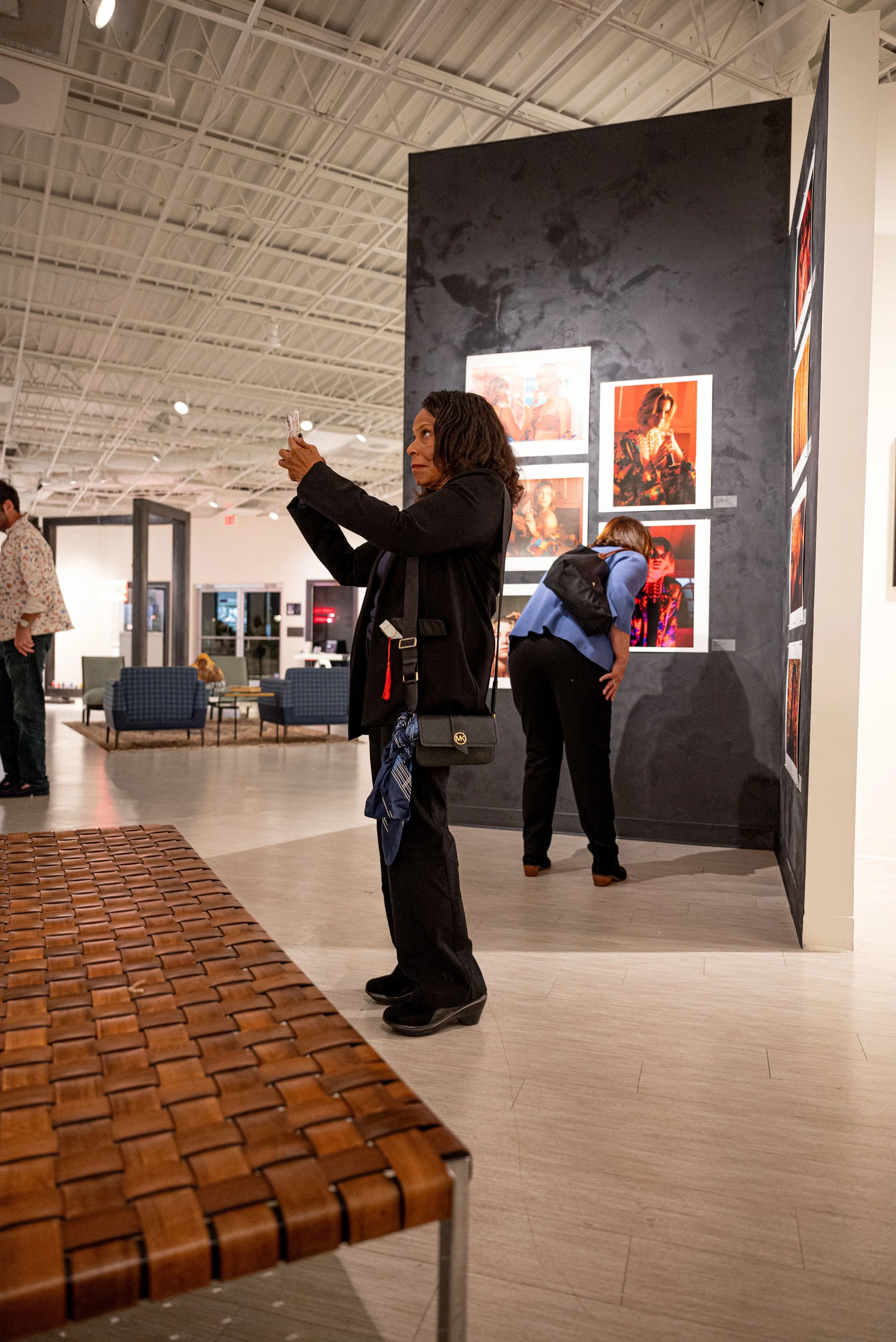 Two women viewing photographs in an art gallery, one taking a picture and the other examining a picture on the wall.