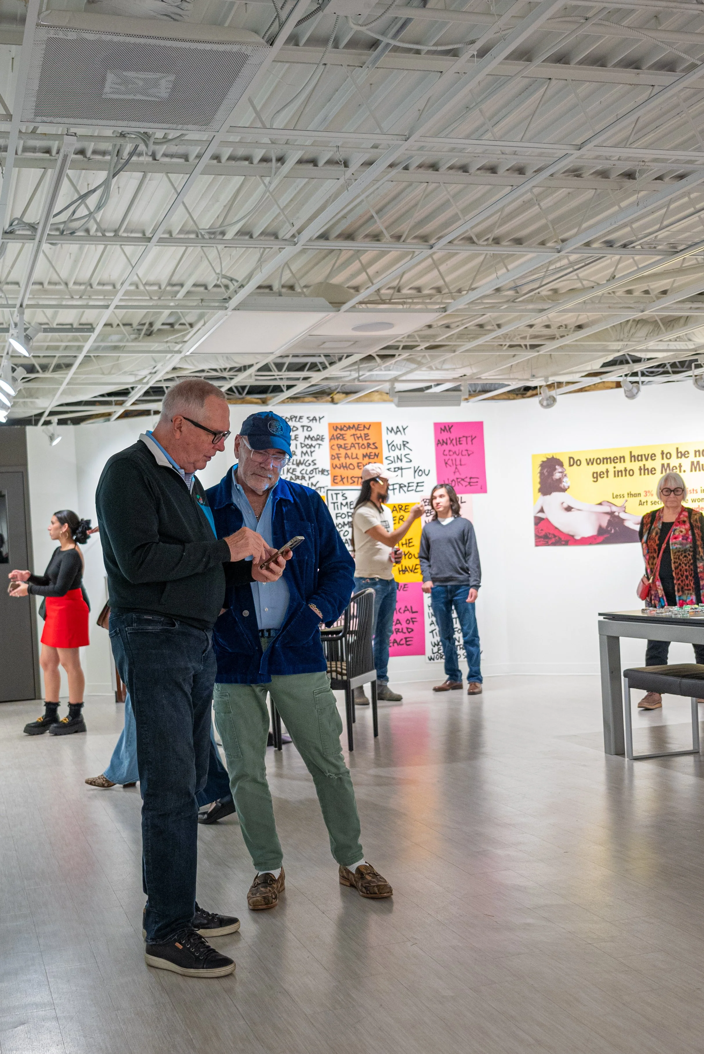 People in an art gallery with colorful signs and posters on the wall, some engaging in conversation and others looking at the art.