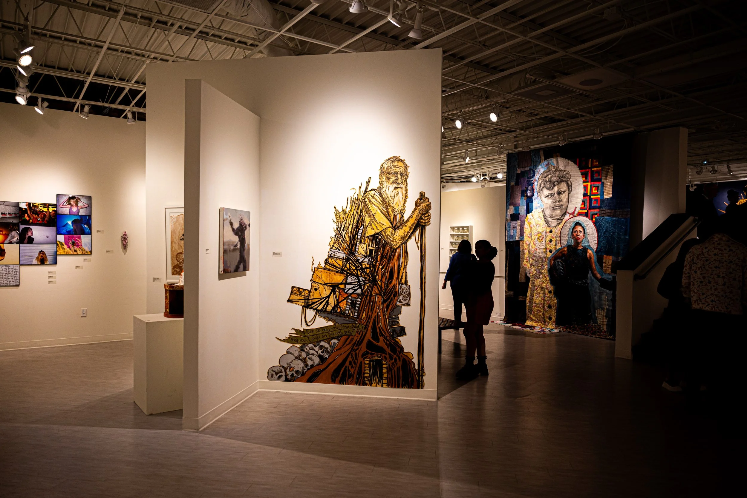 A contemporary art gallery with various murals and artworks displayed on white walls. Prominent in the foreground is a detailed mural of an elderly man with a beard and long hair, holding a stick, painted in a stylized manner with warm tones. In the 