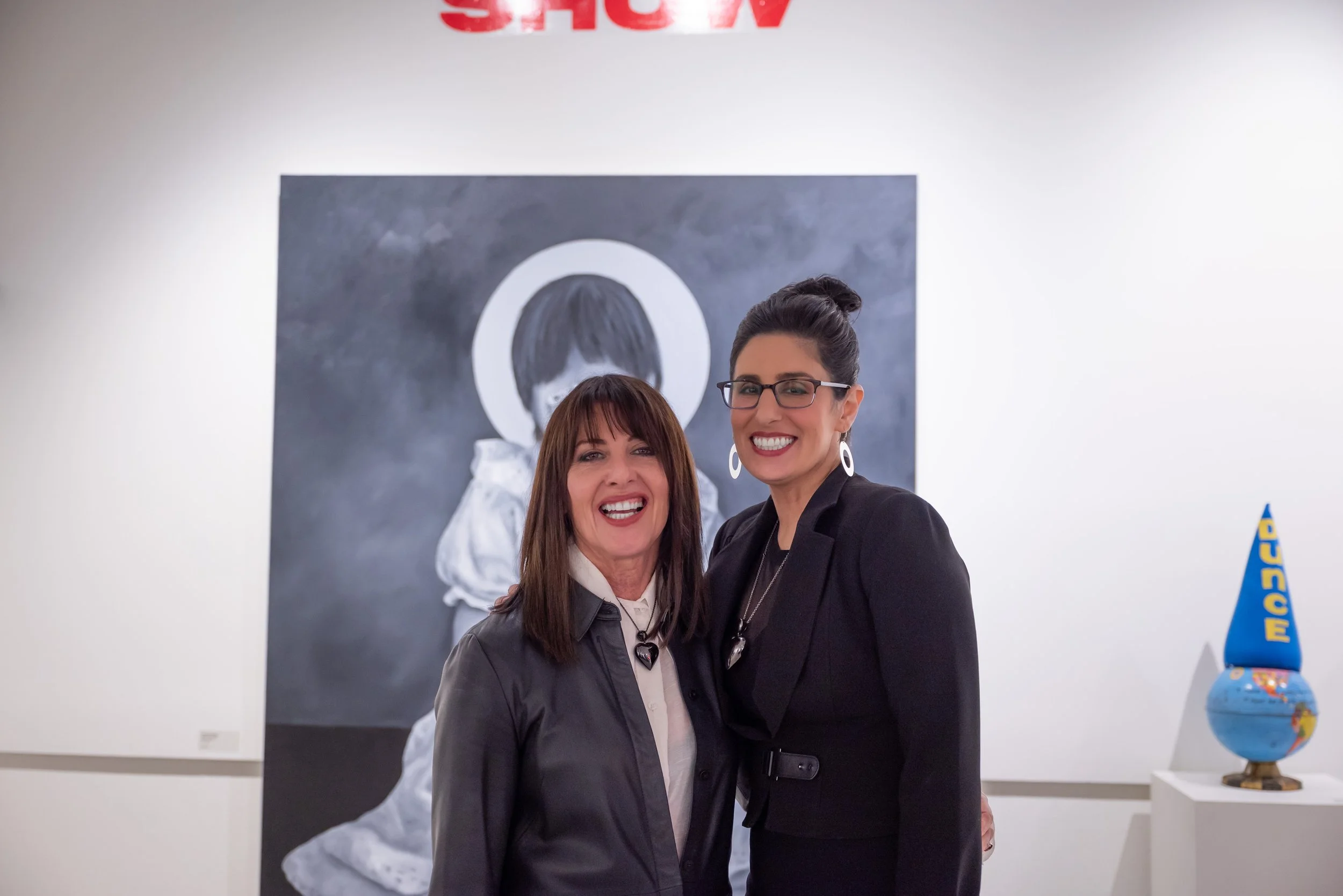 Two women smiling and standing together in an art gallery, with a painting of a person with a bowl haircut in the background and a globe sculpture labeled "Dunce" on a pedestal to the right.
