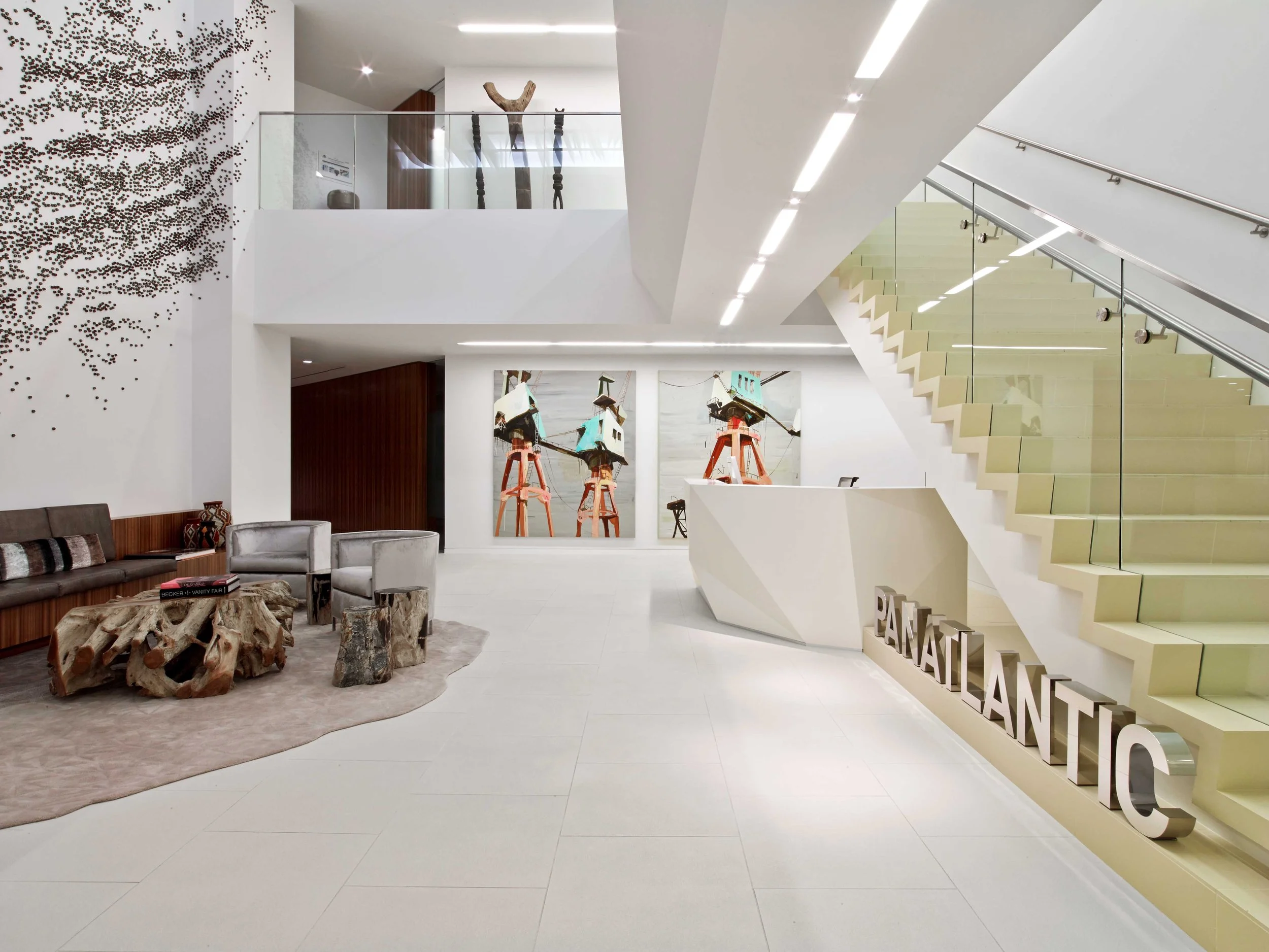 Modern hotel lobby with white walls and staircase, abstract wall art, a sitting area with a rug and unique wooden coffee tables, and the word 'PANATTA' on the counter.