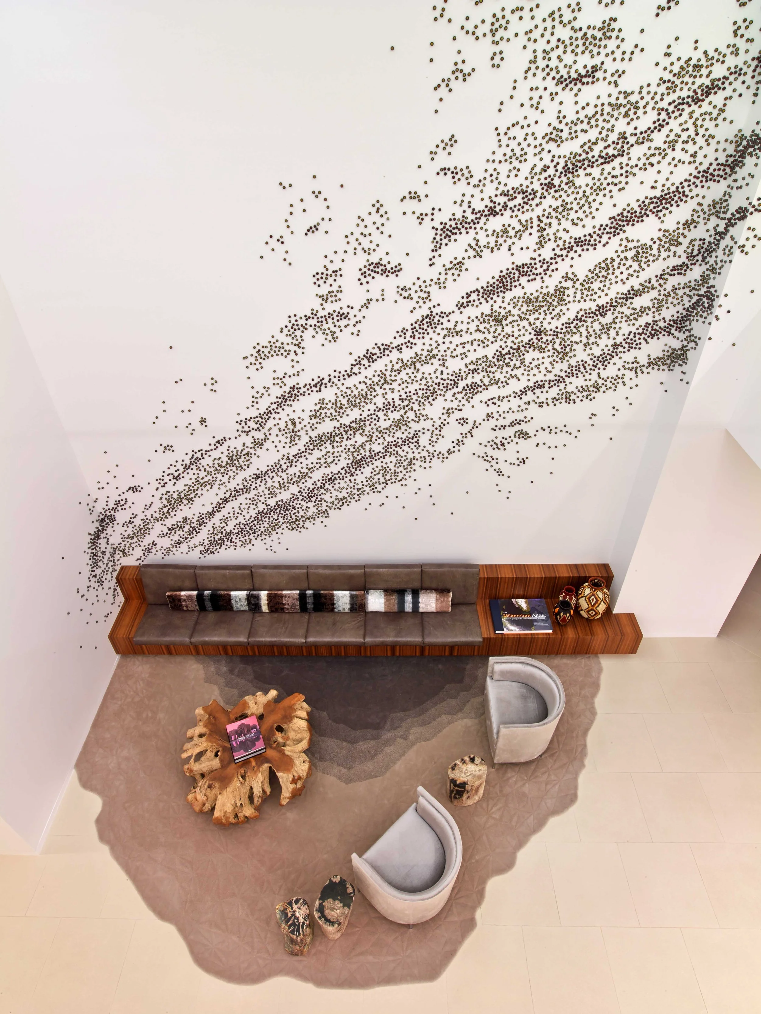 A modern interior with a white wall decorated with black dots creating a swirling pattern. There is a long wooden bench with a brown cushion and decorative pillows, a beige curved chair, and two small table-like seats made of stone or similar materia